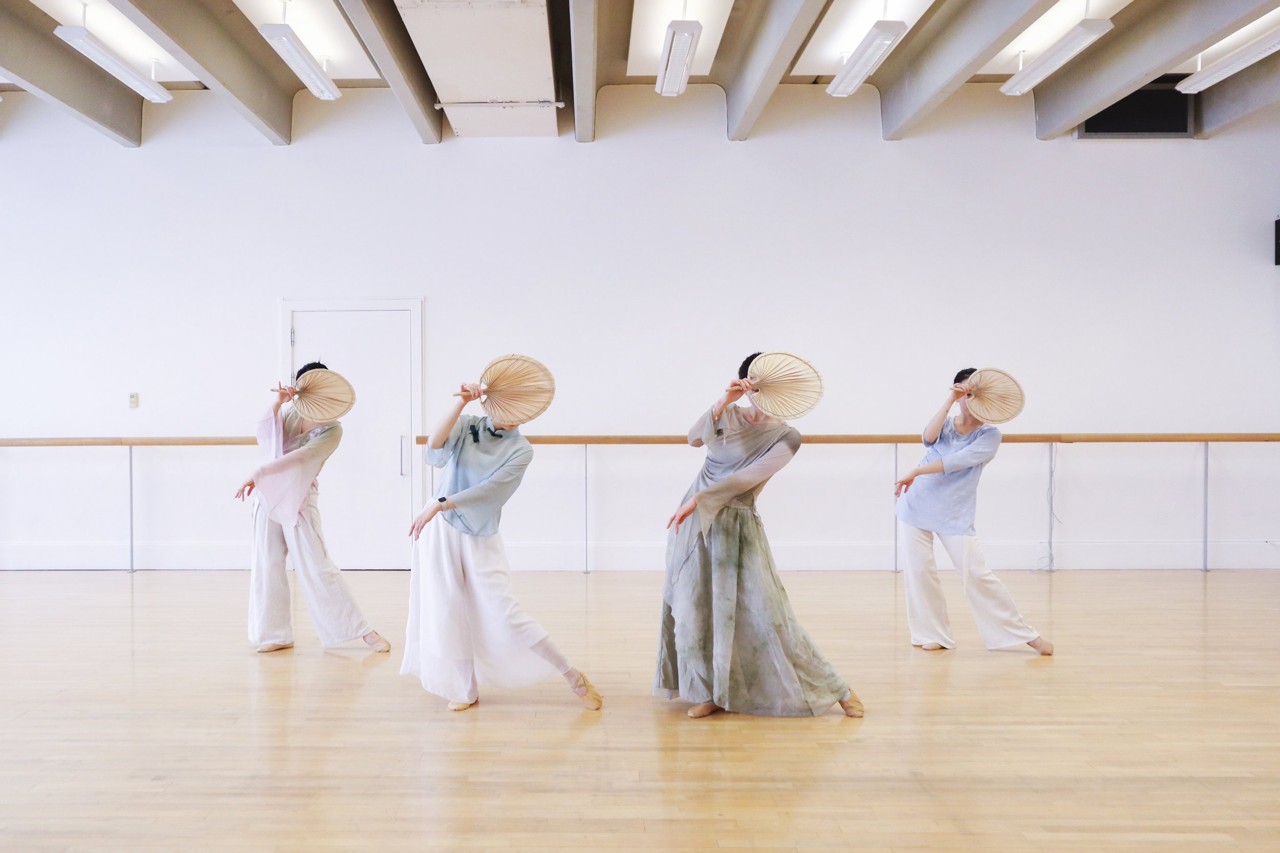 Five women dressed in loose, flowing clothing practicing dance or movement exercises while holding straw fans in a spacious, bright dance studio with a wooden floor and ballet barres.