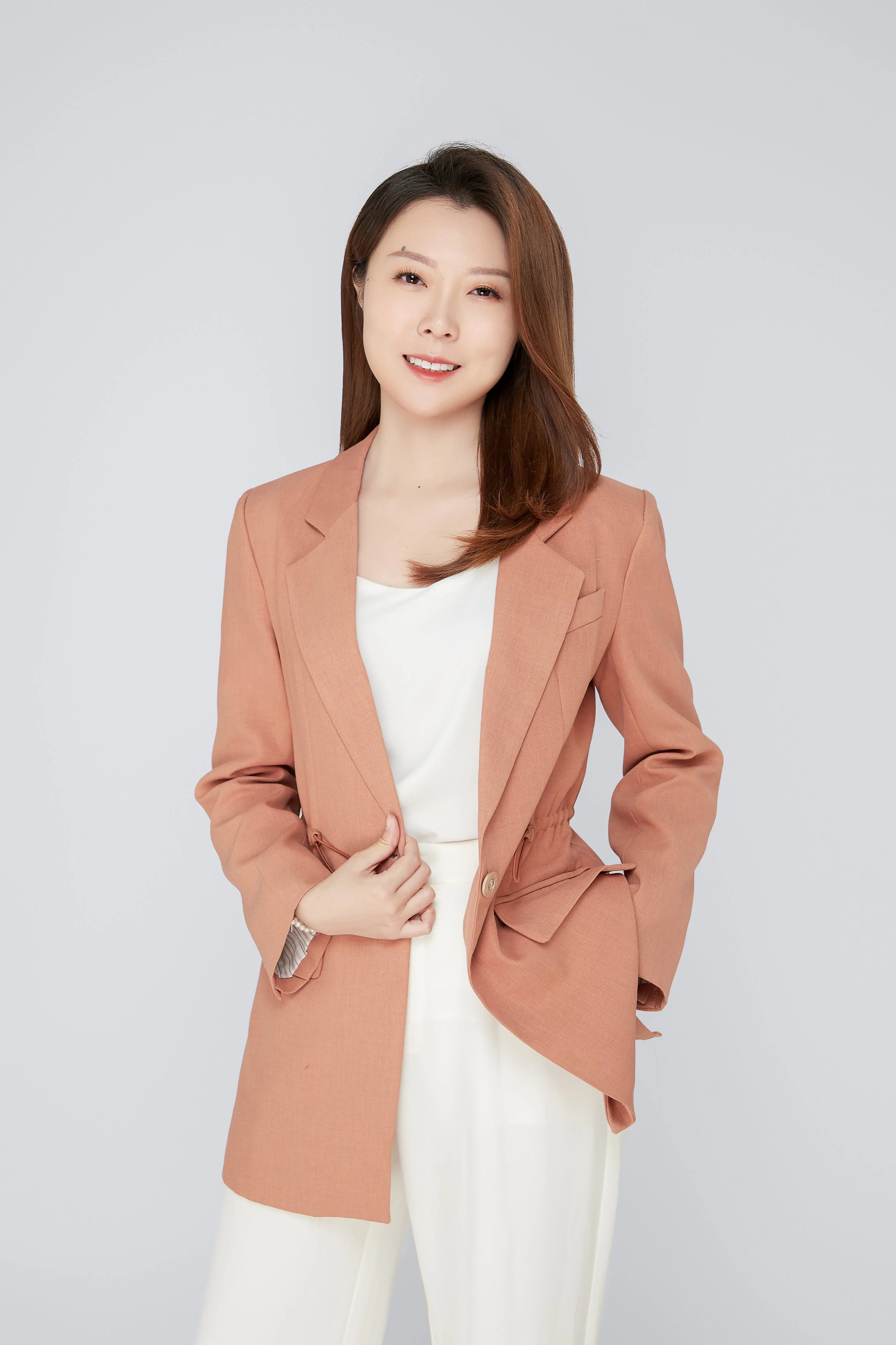 A woman with long brown hair, wearing a salmon-colored blazer over a white dress, standing against a plain gray background, smiling and looking at the camera.