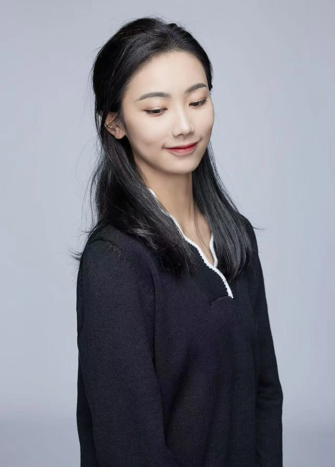 A woman with long black hair and light complexion, wearing a black jacket with a white collar, looking down with a slight smile, set against a plain gray background.