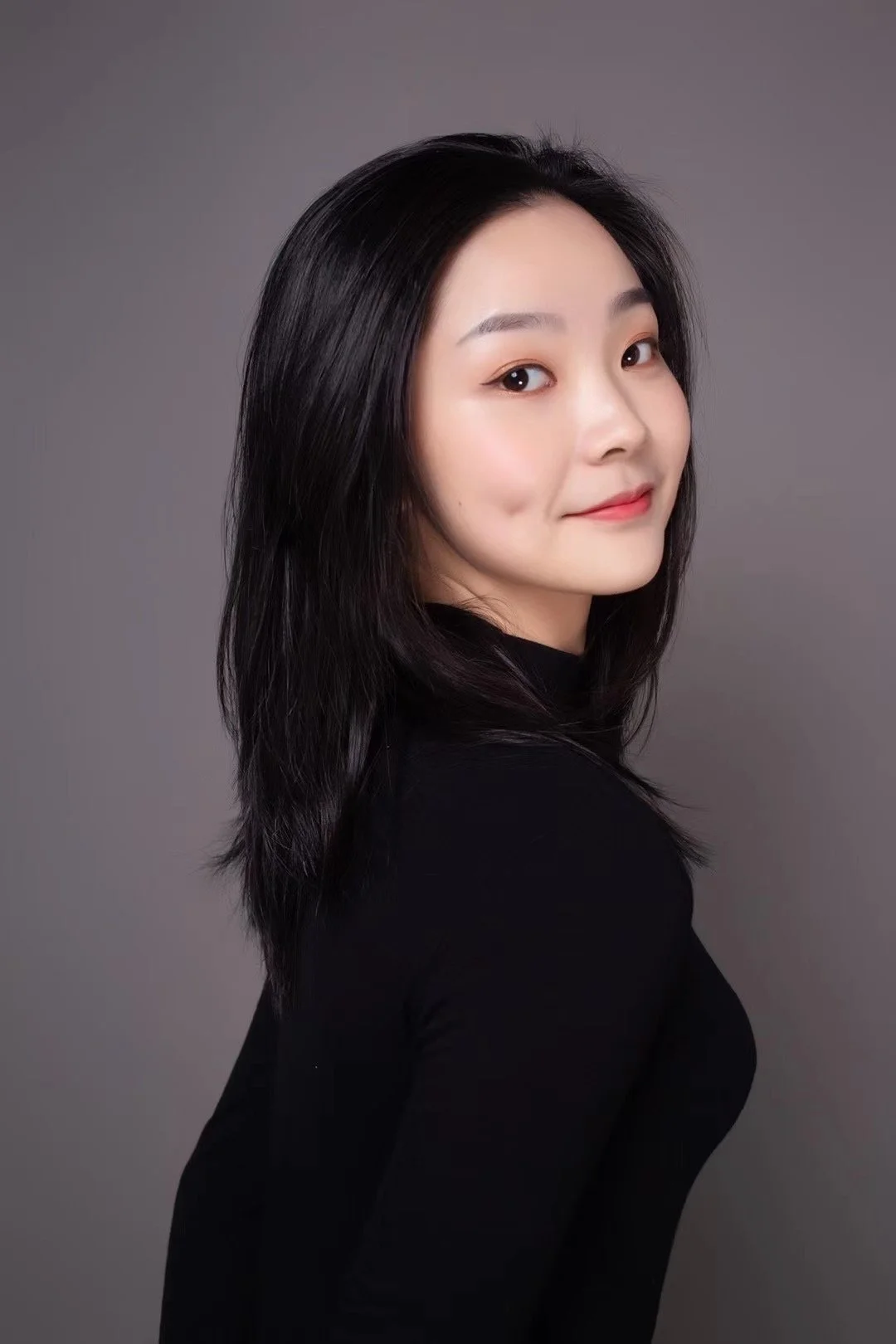 A woman with black hair and fair skin, wearing a black top, smiling slightly, against a solid gray background.