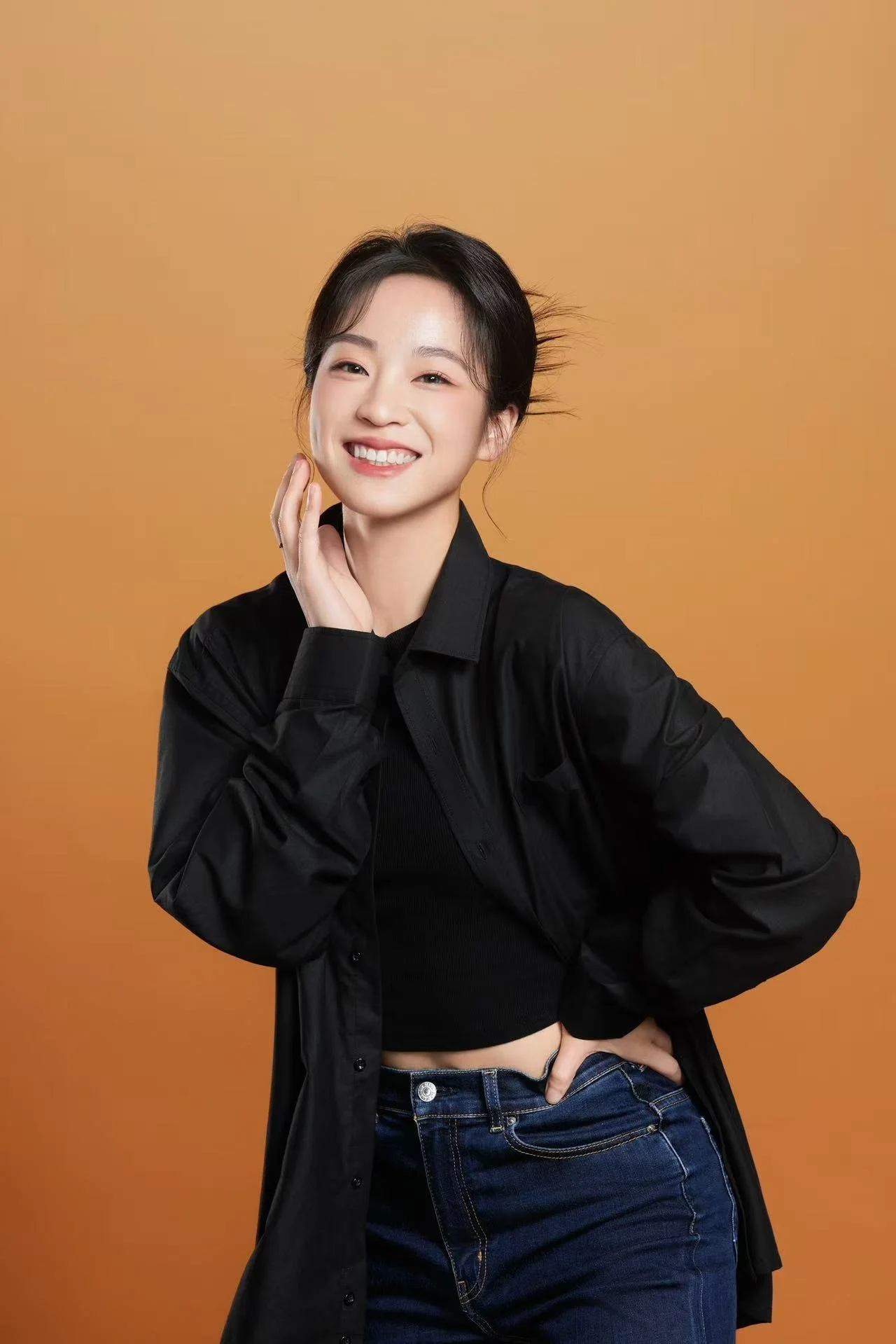 A young woman with short dark hair, smiling, wearing a black shirt and jacket, with one hand on her face and the other on her waist, standing against an orange background.