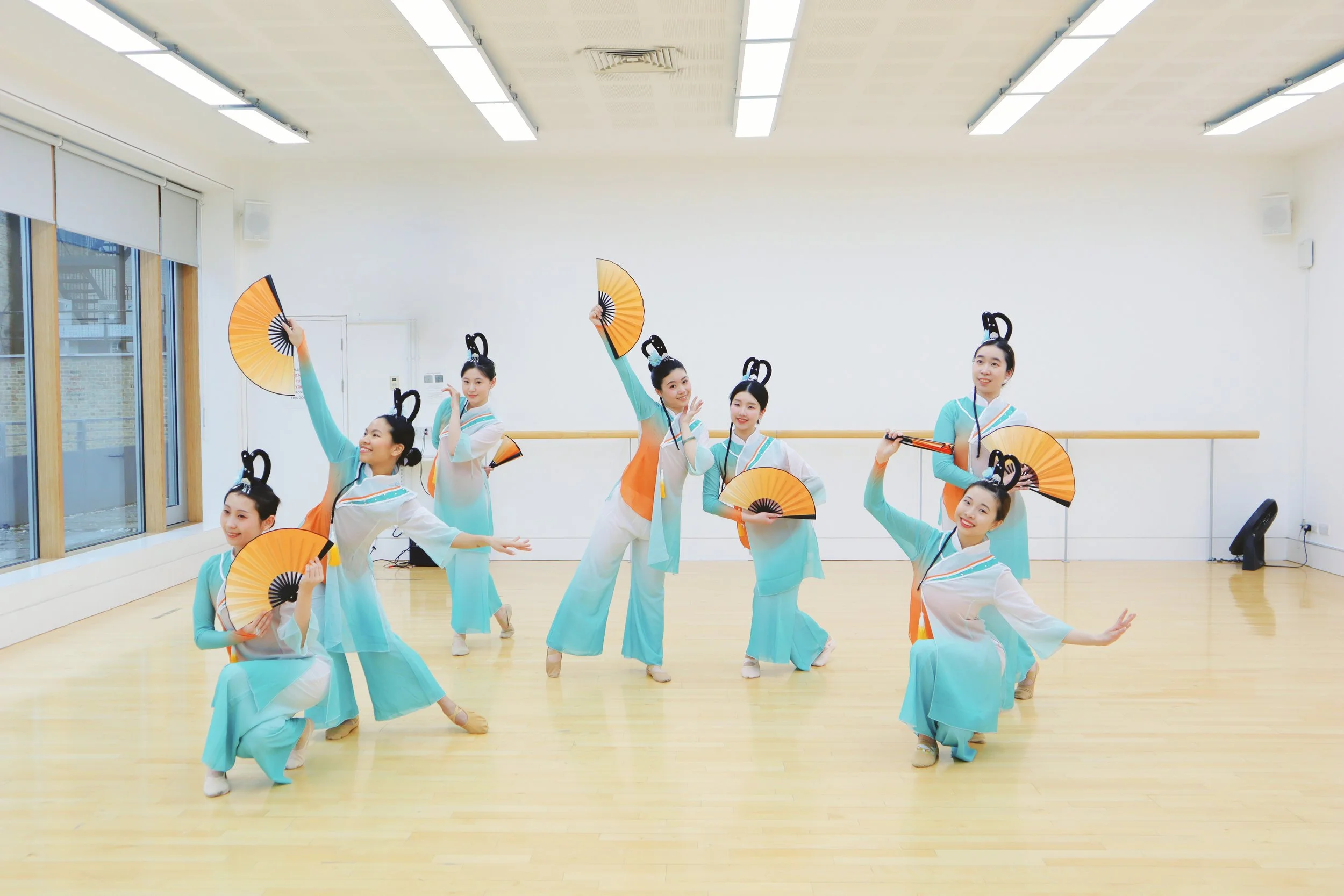 Group of women dressed in traditional Chinese attire performing a fan dance in a dance studio.