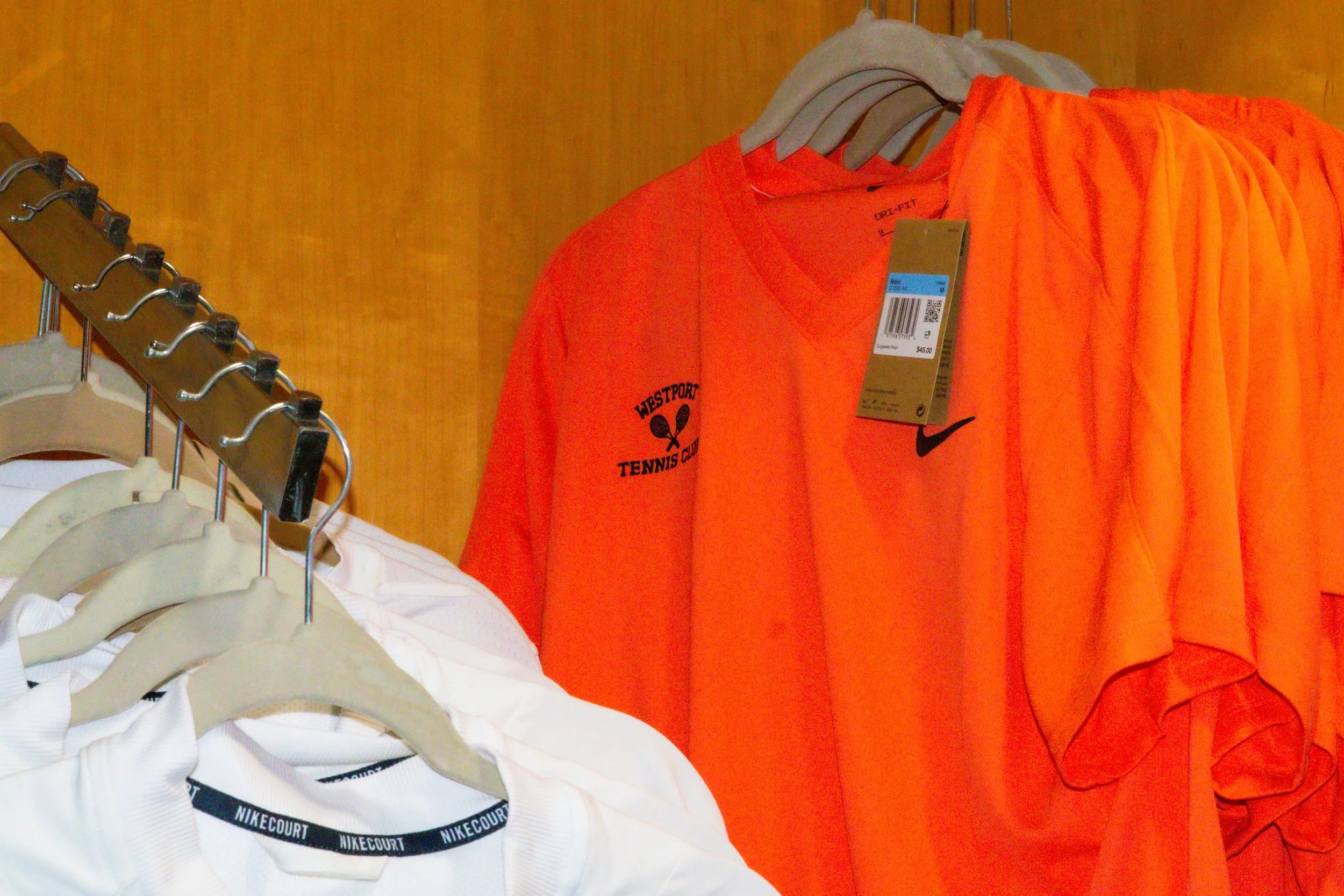 Orange Nike tennis shirt with Westport Tennis Club logo displayed alongside white Nike apparel in the pro shop.