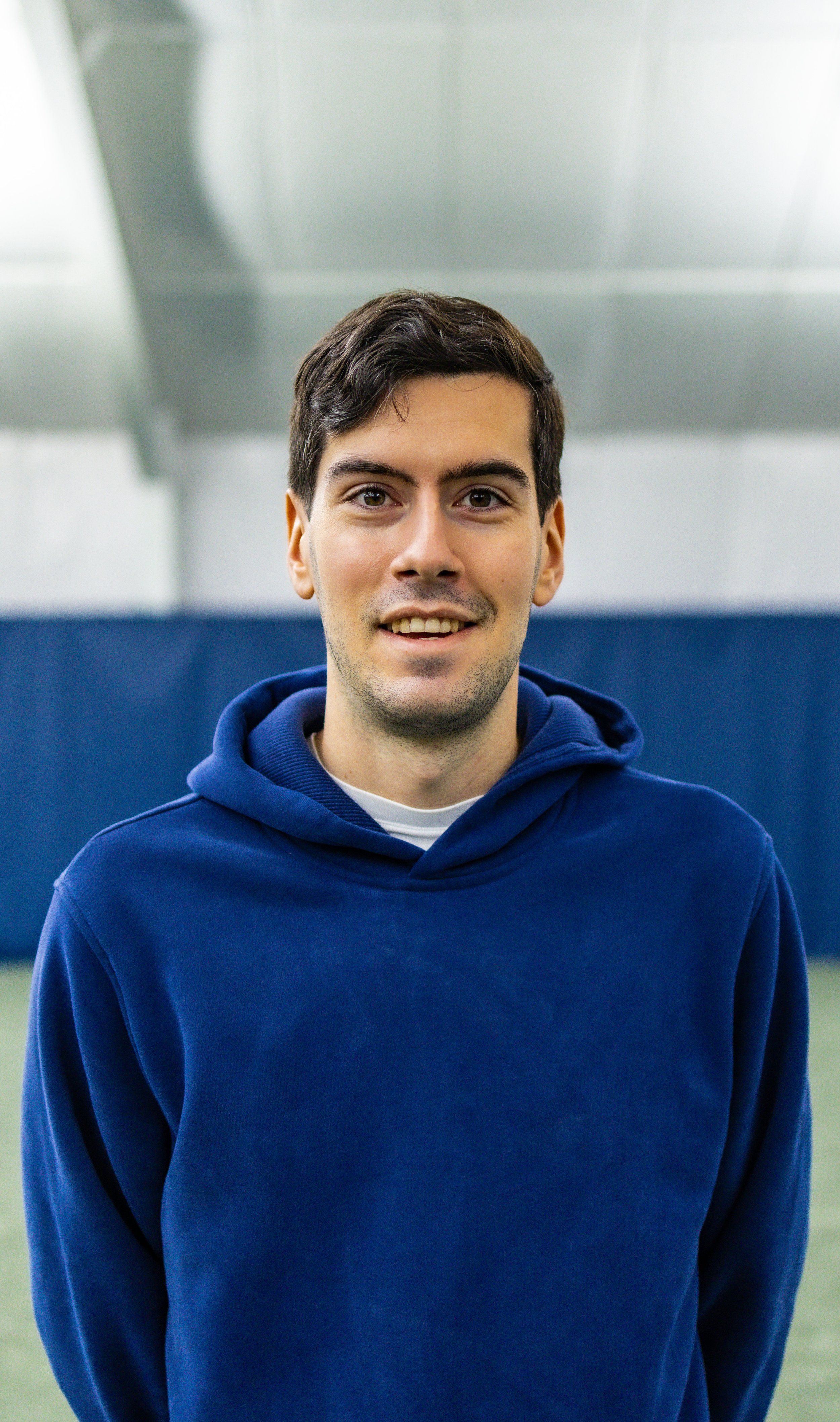Young man standing indoors at Westport Tennis Club, wearing a blue hoodie with a blurred indoor tennis background.