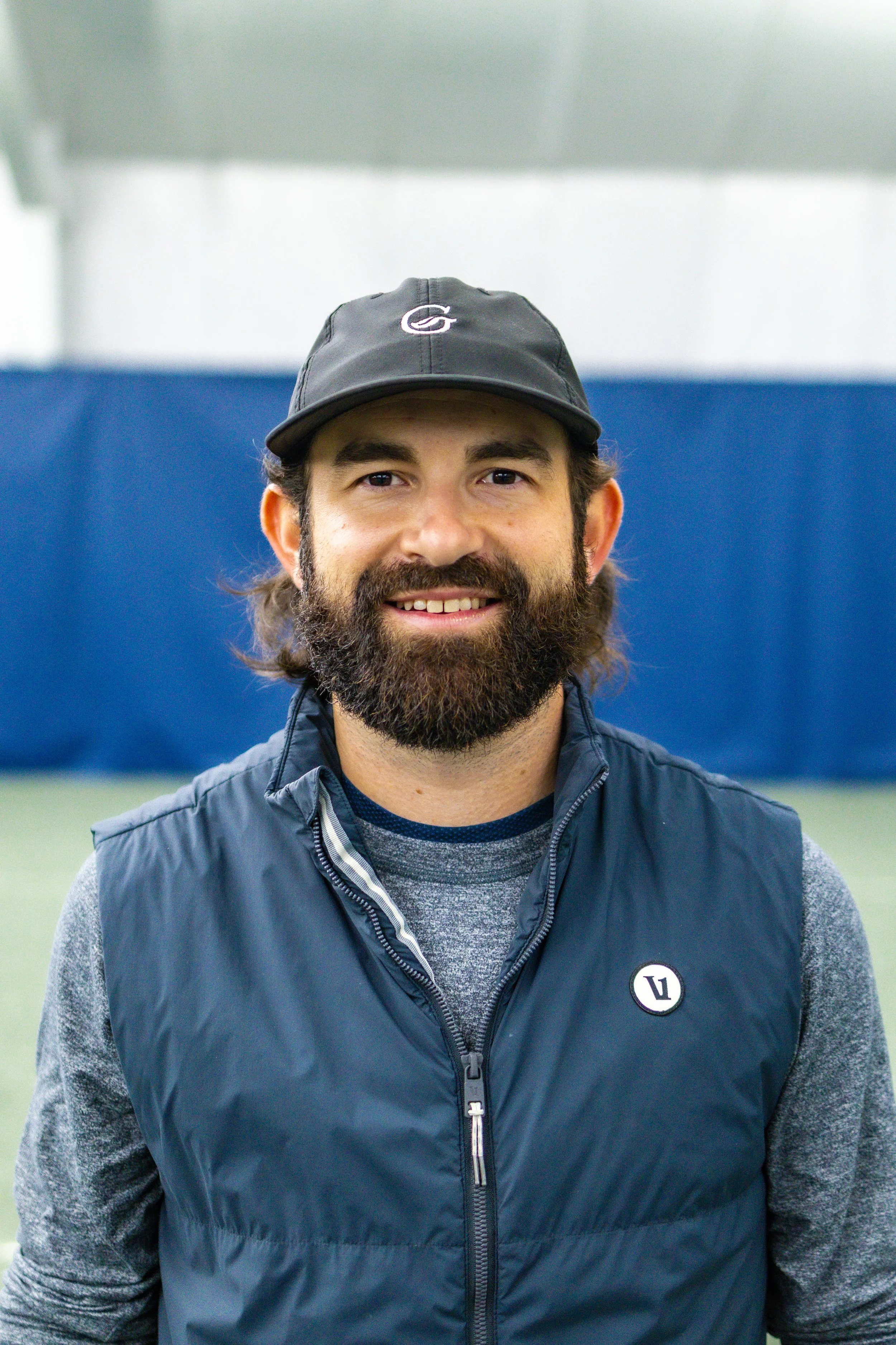 Smiling tennis instructor with a beard, wearing a black cap and blue vest, standing inside Westport Tennis Club.