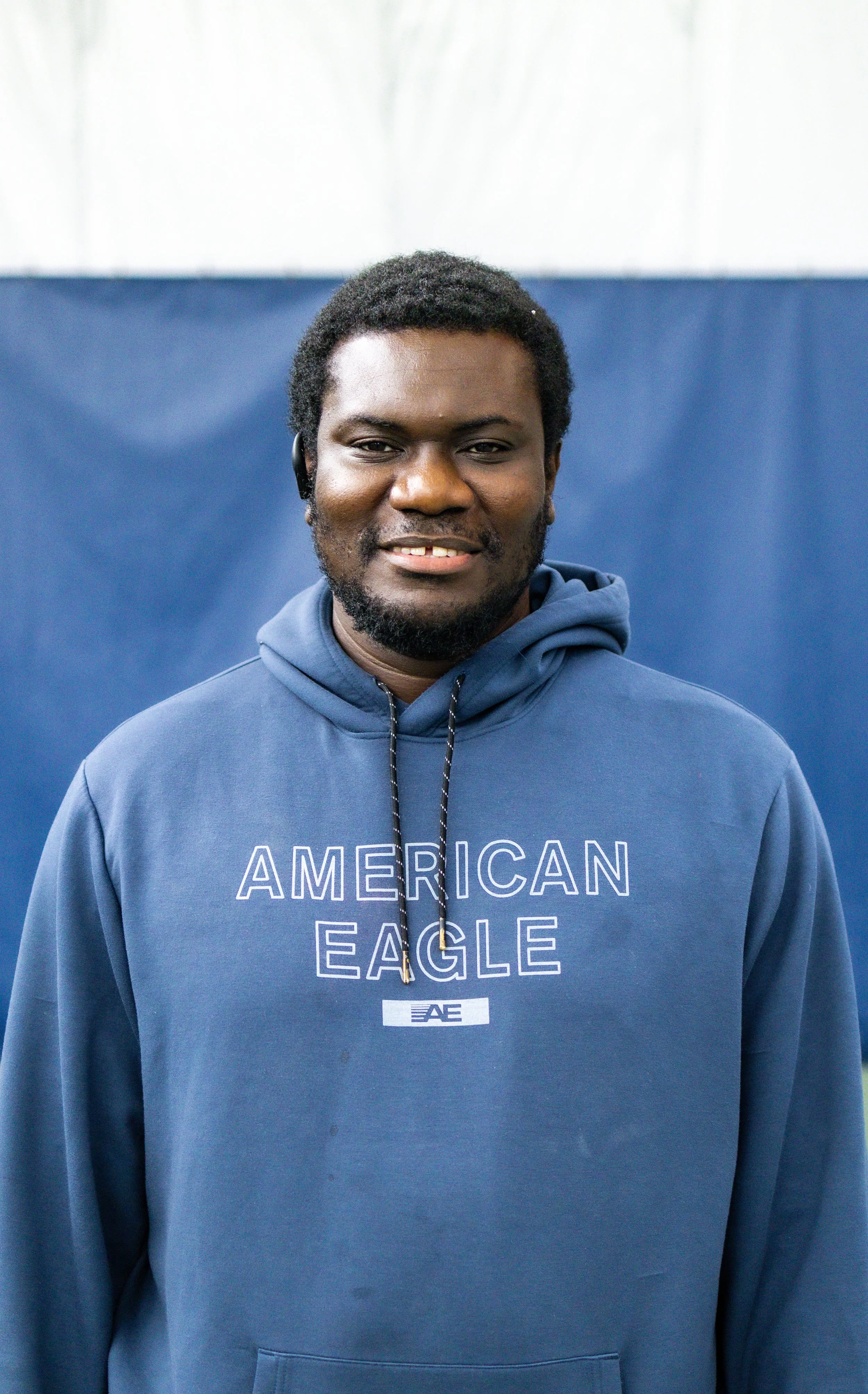 Young male staff member smiling at Westport Tennis Club, wearing a blue American Eagle hoodie in front of a blue and white background.