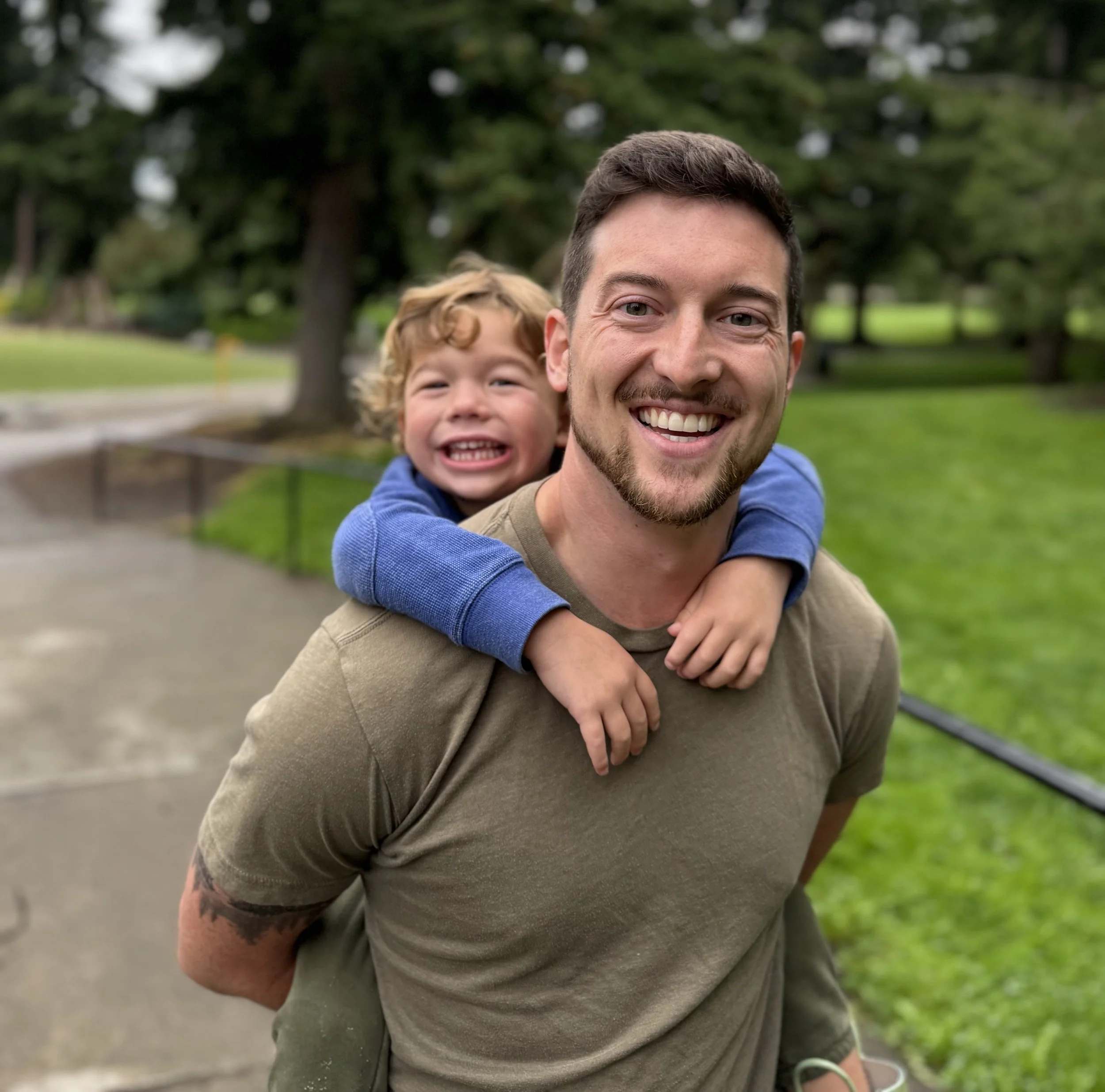 A smiling man giving a piggyback ride to a young boy in a park with green grass and trees.