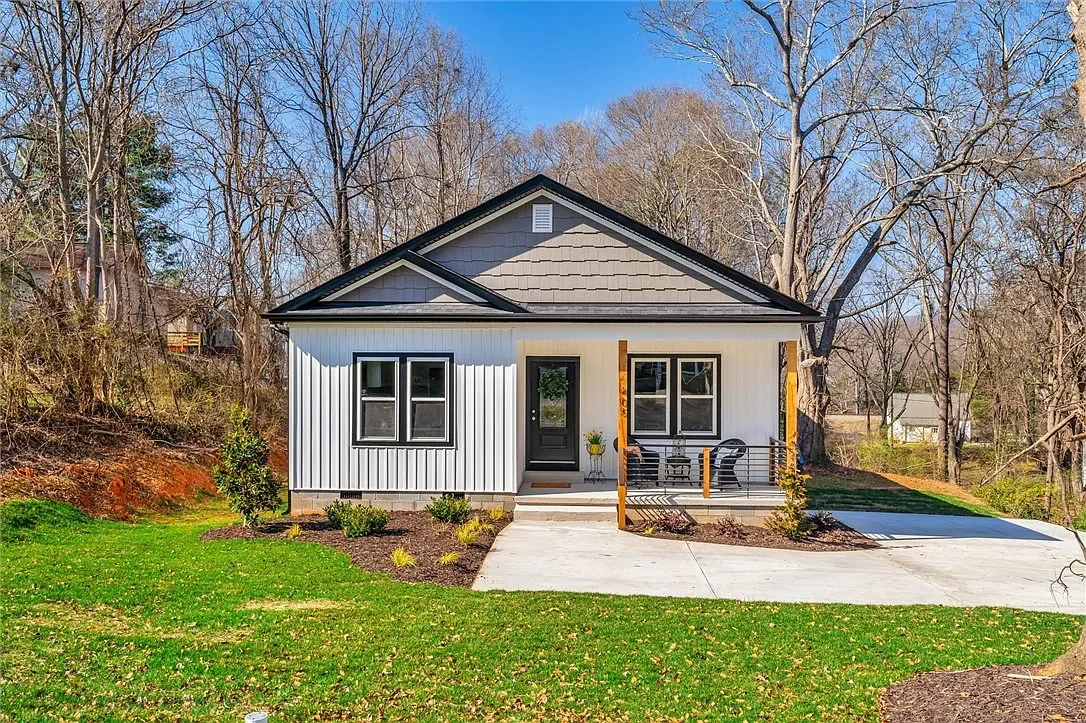 Newly constructed small house with gray shingle roof, white siding, black front door, and small porch with black railing in a suburban neighborhood with leafless trees.