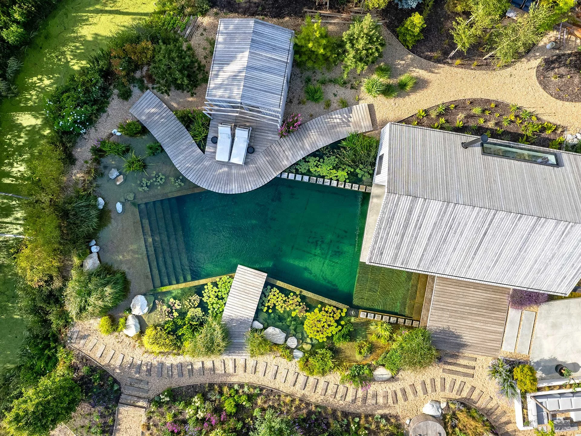 Aerial view of a backyard with a swimming pool, wooden deck, garden beds, and surrounding greenery and trees. Natural Swimming Pool design