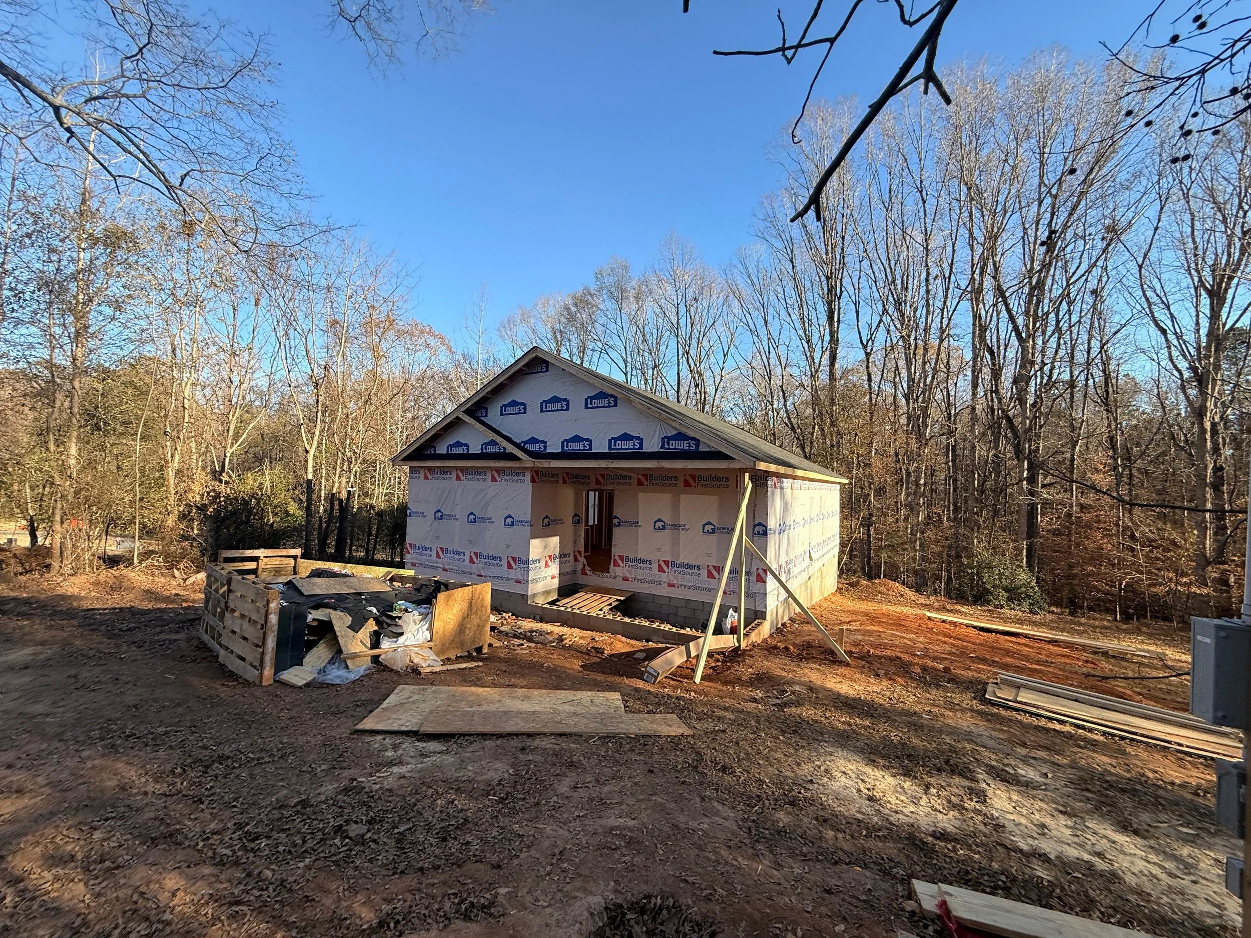 Construction site with a small house being built, wrapped in Builders Building Wraps, surrounded by trees with no leaves, under a clear blue sky. Woodruff, SC Framing of a New Home Construction