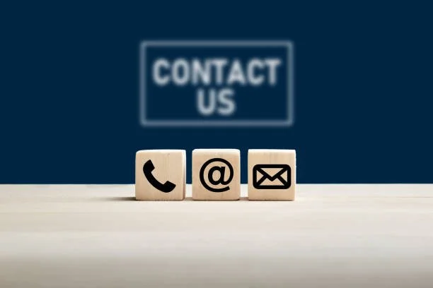 Three wooden blocks with icons of a phone, an at symbol, and an envelope, in front of a sign that says "Contact Us" on a blue background.