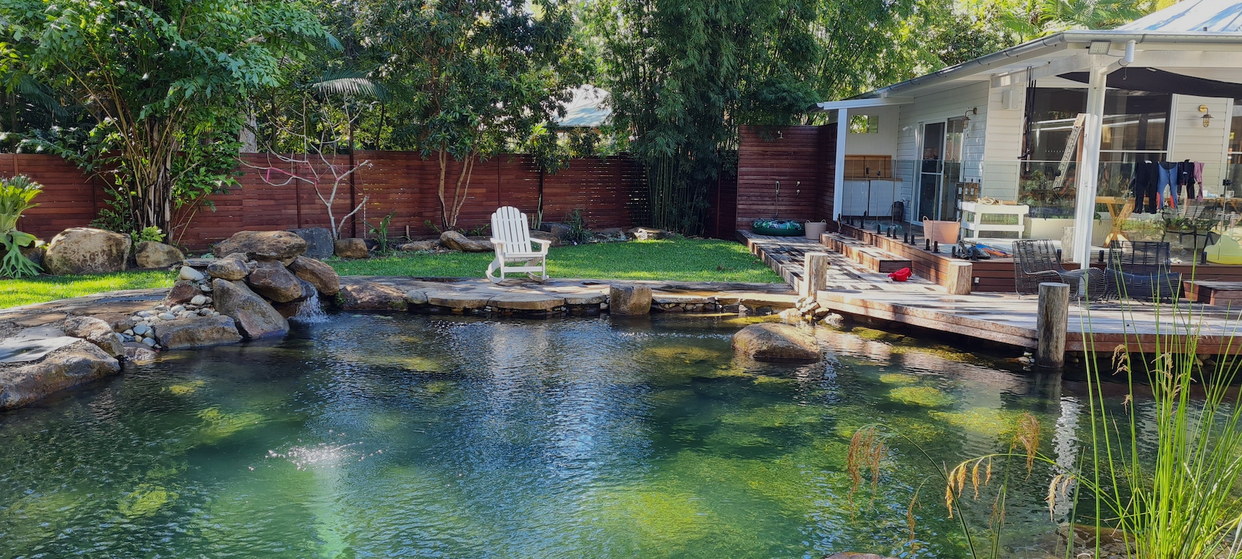 Backyard with pond, rocks, trees, and a wooden deck with outdoor furniture and house in the background.