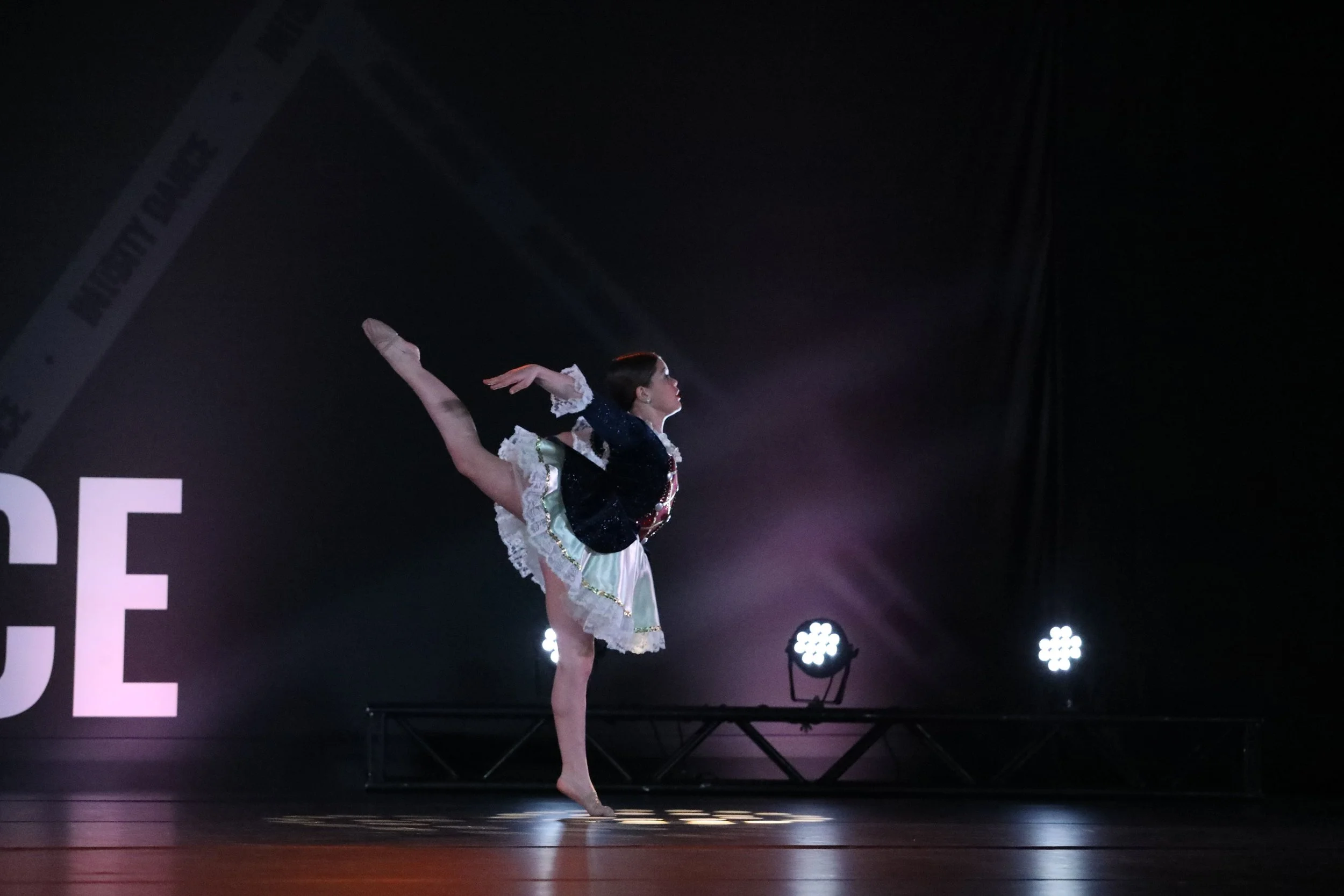A female dancer performing on stage, balancing on one foot with the other leg extended behind her, wearing a black and white costume with frills, against a dark background with stage lights.
