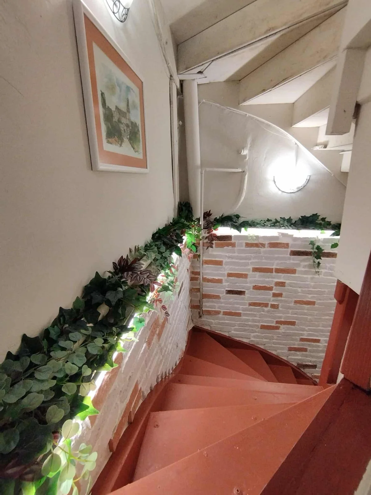 Interior view of a spiral staircase with brick walls and a wooden handrail, decorated with a string of green ivy plants. There is a framed picture on the wall, and a round wall light illuminating the staircase.