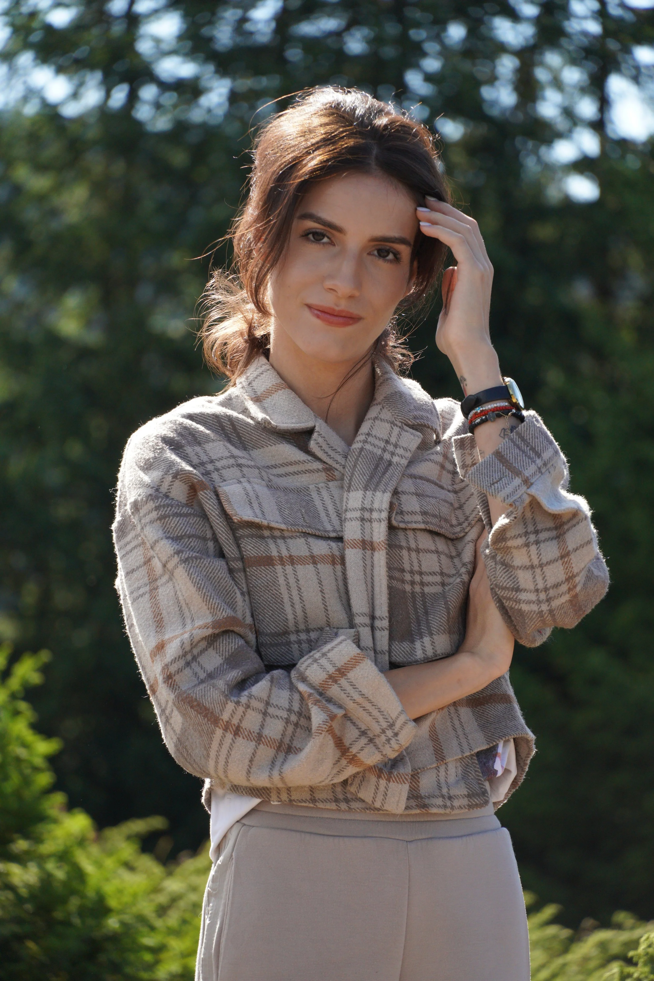 A woman with long brown hair, wearing a beige plaid jacket, smiling outdoors on a sunny day with trees in the background.
