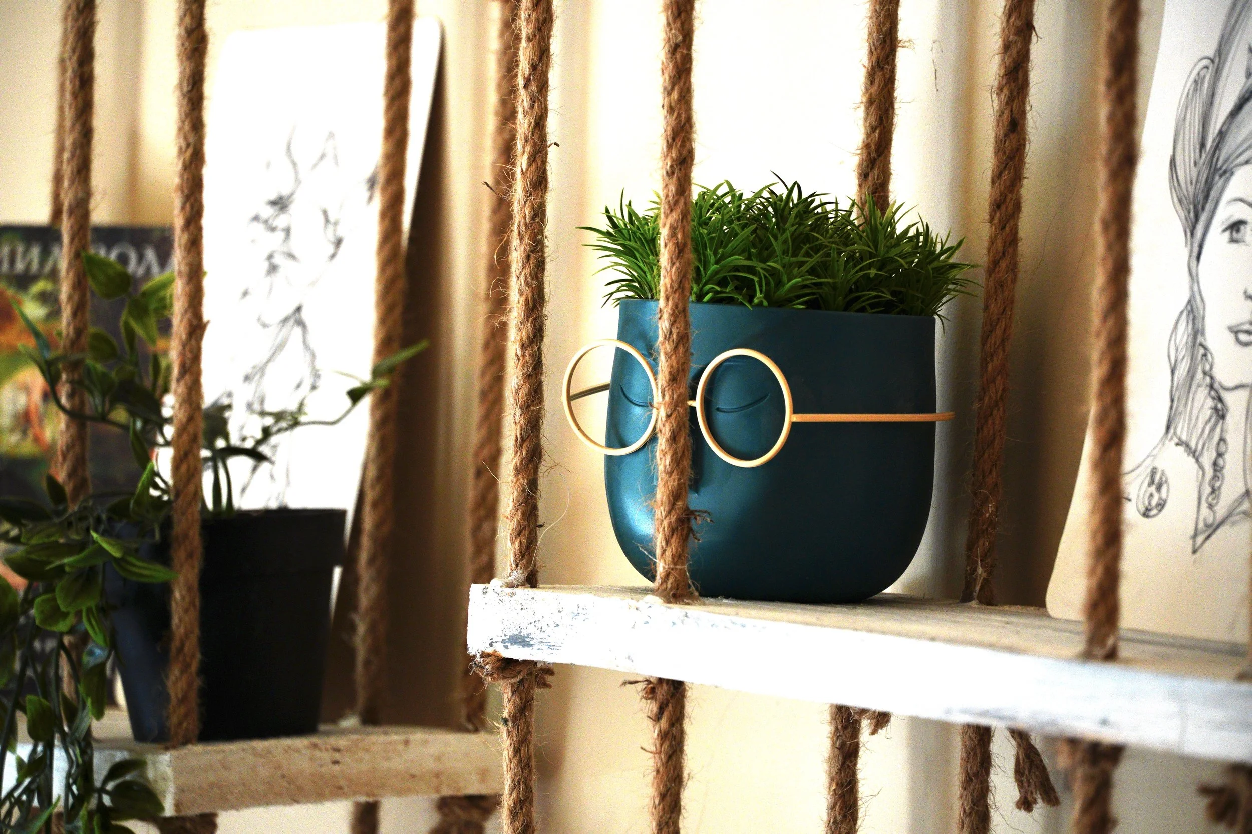 A blue planter with green plants on a white wooden shelf, seen through brown ropes, with a pair of gold glasses hanging on the side of the planter.