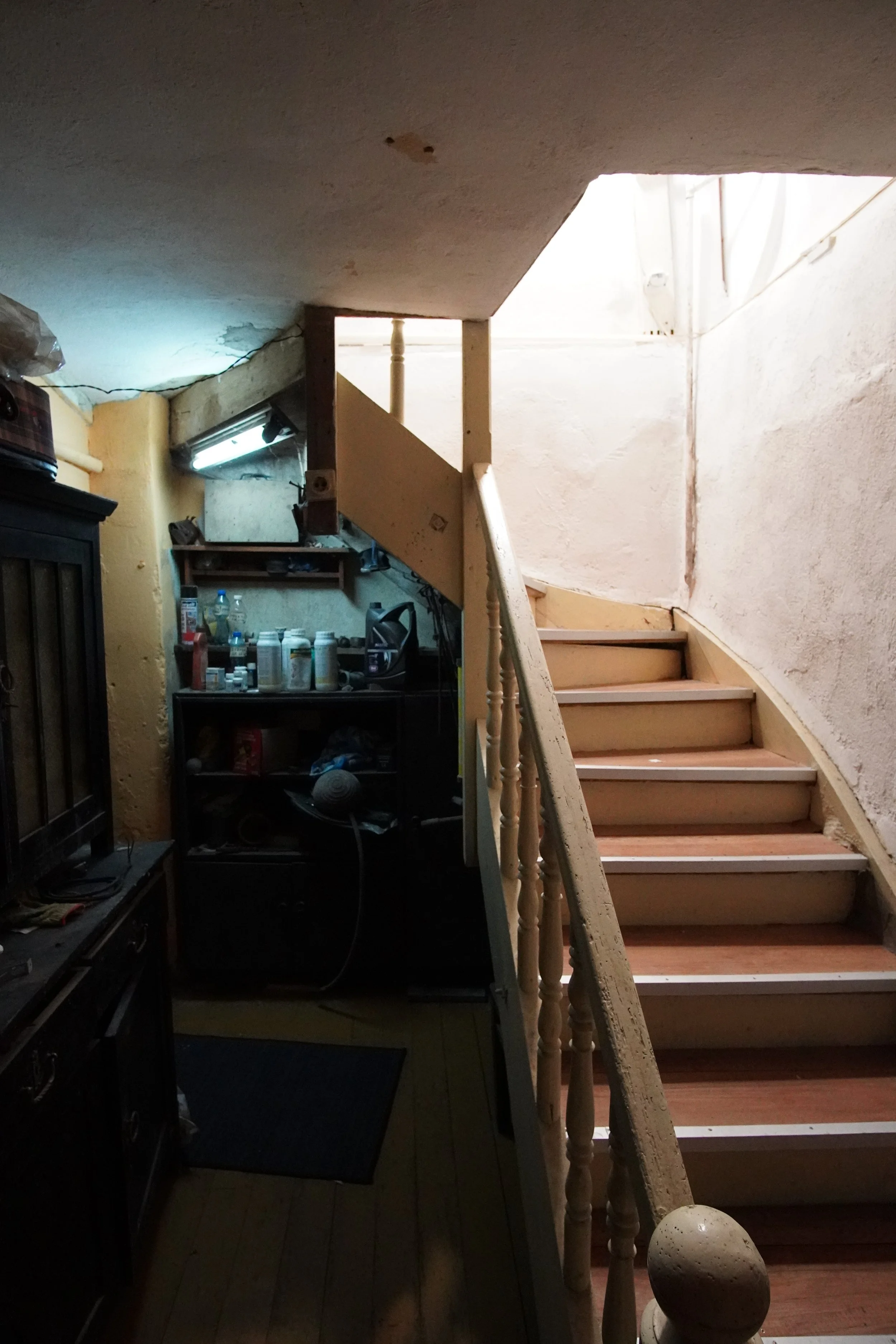 Interior of a small, dimly lit room with a wooden staircase leading to an upper level, cluttered with household items and shelves.