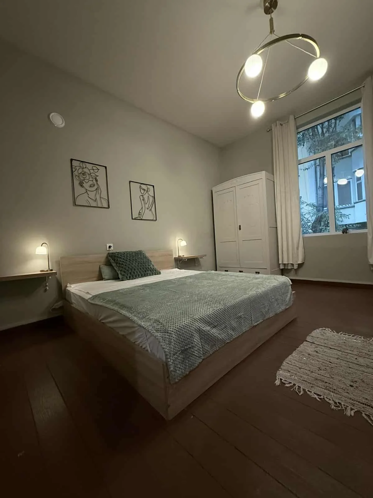 A cozy bedroom with a large bed, green textured bedding, and a matching green throw pillow. There are two wall-mounted reading lamps on either side of the bed. Above the bed are minimalist line art pictures. To the right, a large window with white cu