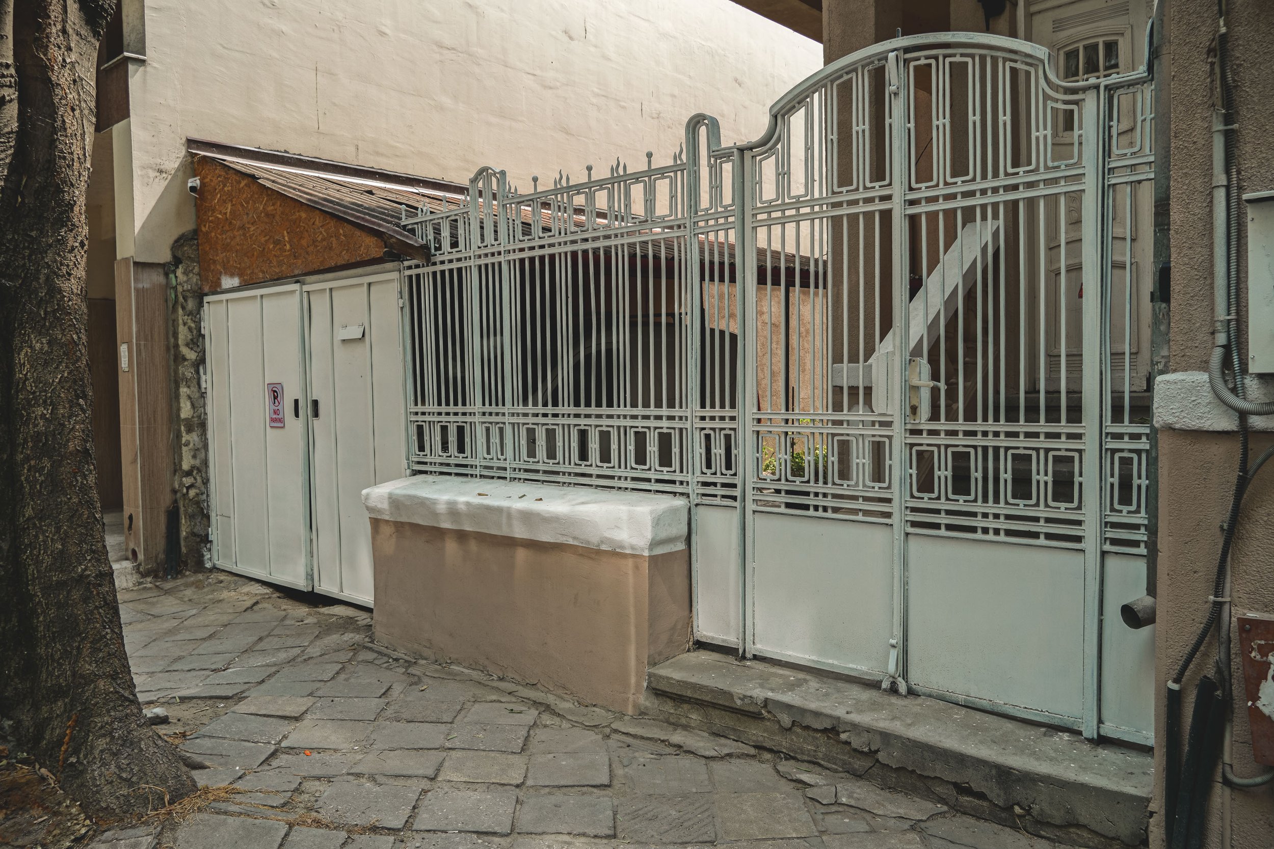 A metal gate with a staircase behind it, located outside a building with stone and stucco walls, on a cobblestone sidewalk near a tree.
