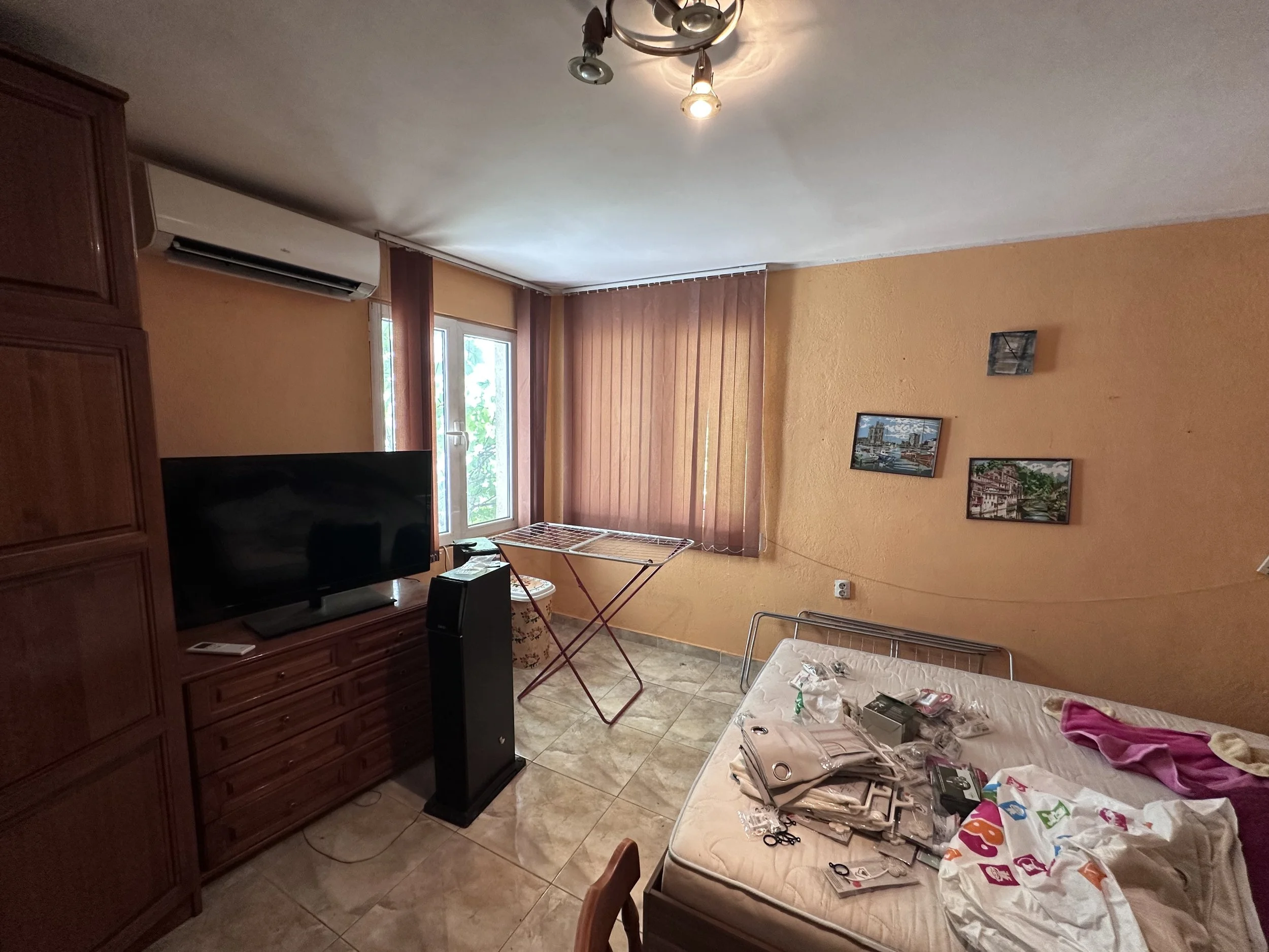 A cluttered bedroom with a bed covered in various items, a flat-screen TV on a wooden cabinet, a window with vertical blinds, a drying rack, a wall air conditioning unit, and framed pictures on the orange wall.