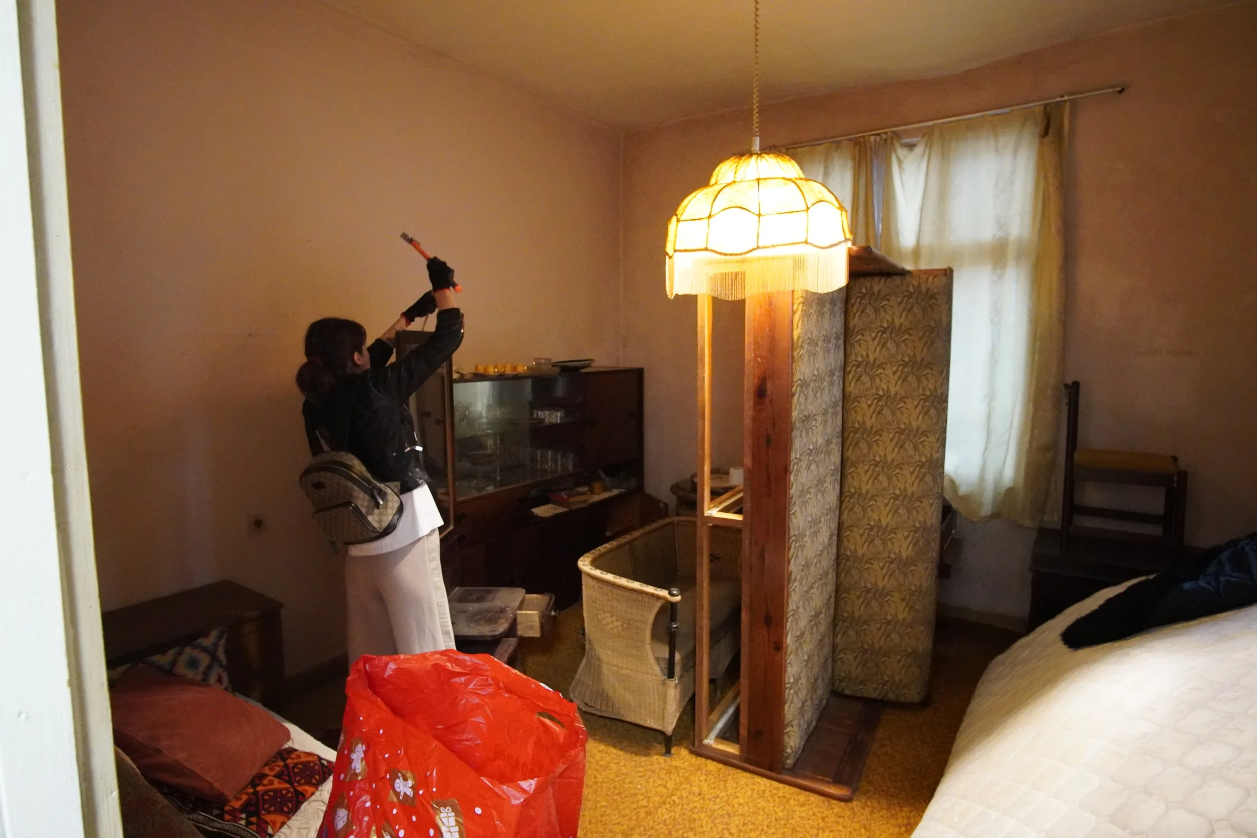 A woman with a backpack and black jacket is holding a baseball bat, standing in a cluttered room with two beds, a screen dividing space, a lamp hanging from the ceiling, and a window with curtains.