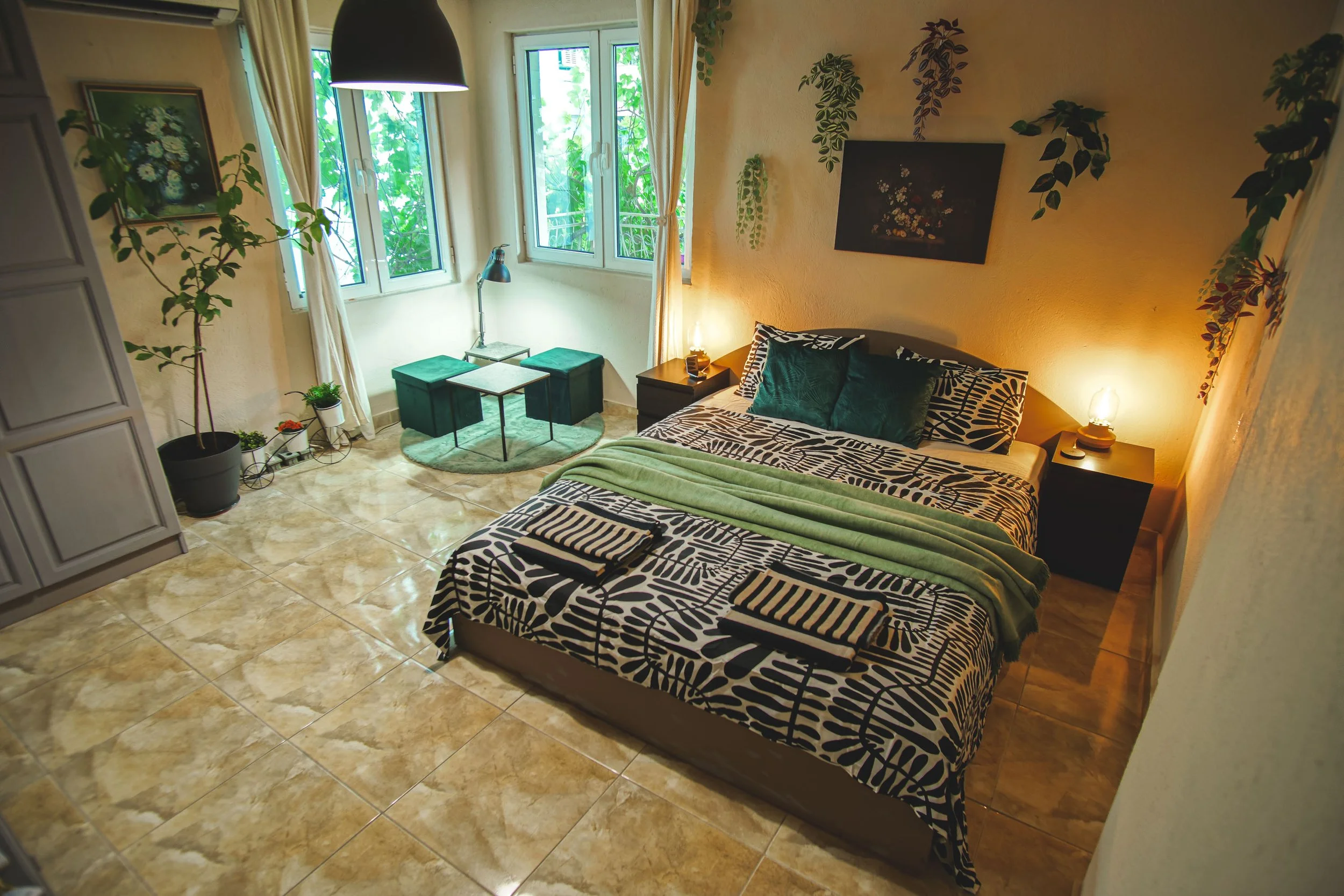 A cozy bedroom with a bed featuring a black and white patterned bedspread and dark green pillows, flanked by two black nightstands with lamps. There are three folded towels on the bed. The room has a window with white curtains, several potted plants,