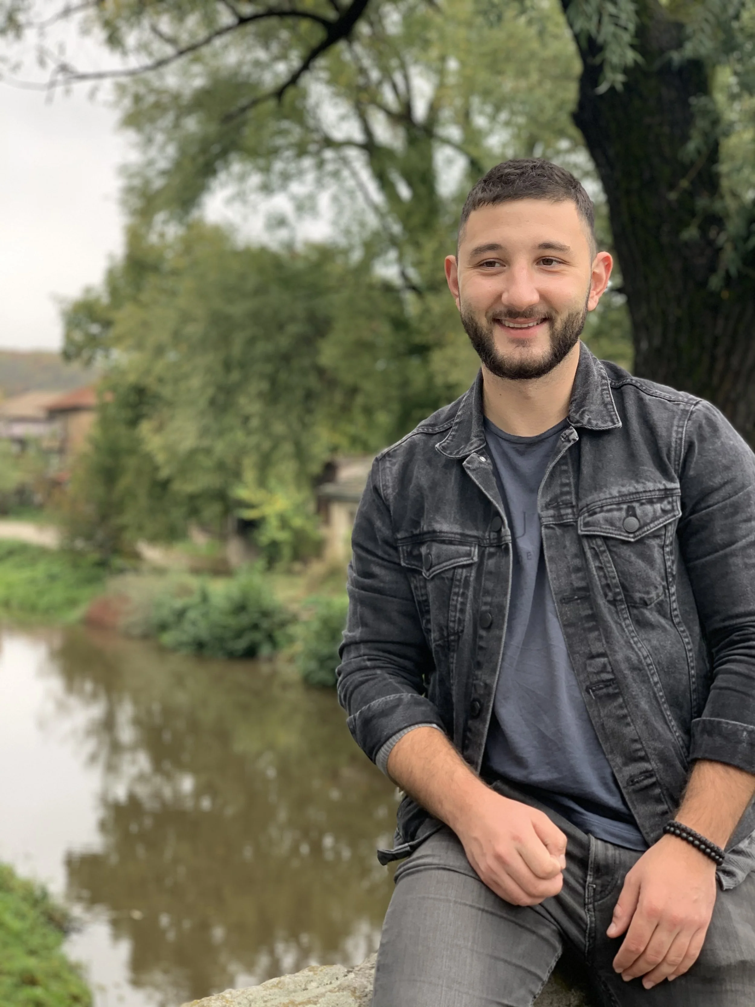 A smiling young man in a black denim jacket and gray pants posing outdoors near a river with lush green trees in the background.