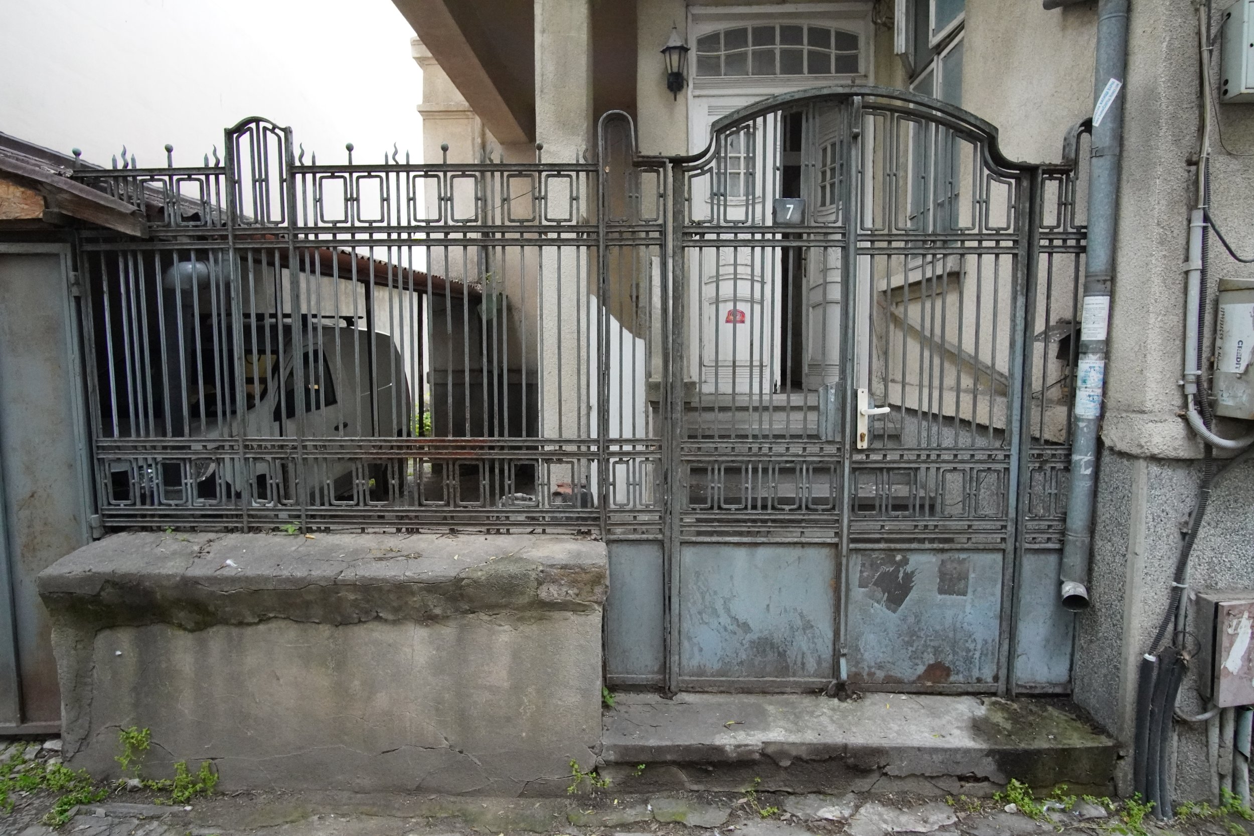 A metal gate with a locked handle, leading to a staircase and the entrance of a building. The gate is situated on a cracked concrete platform with small green plants growing at its edge.