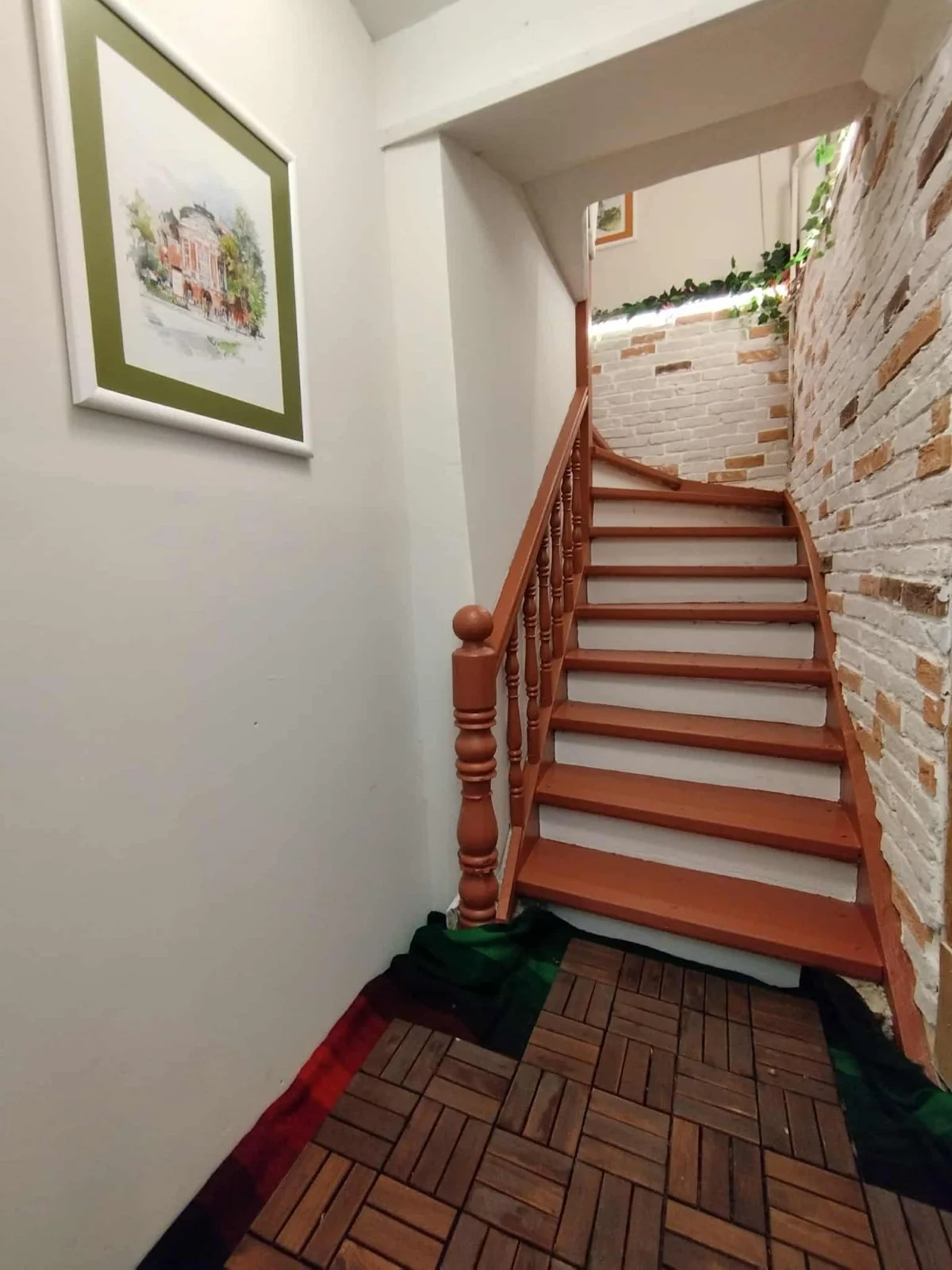 An interior staircase with wooden railing and steps, white walls, a brick accent wall on the right, a framed watercolor picture on the left, and green plants at the top, with a patterned floor mat at the bottom.