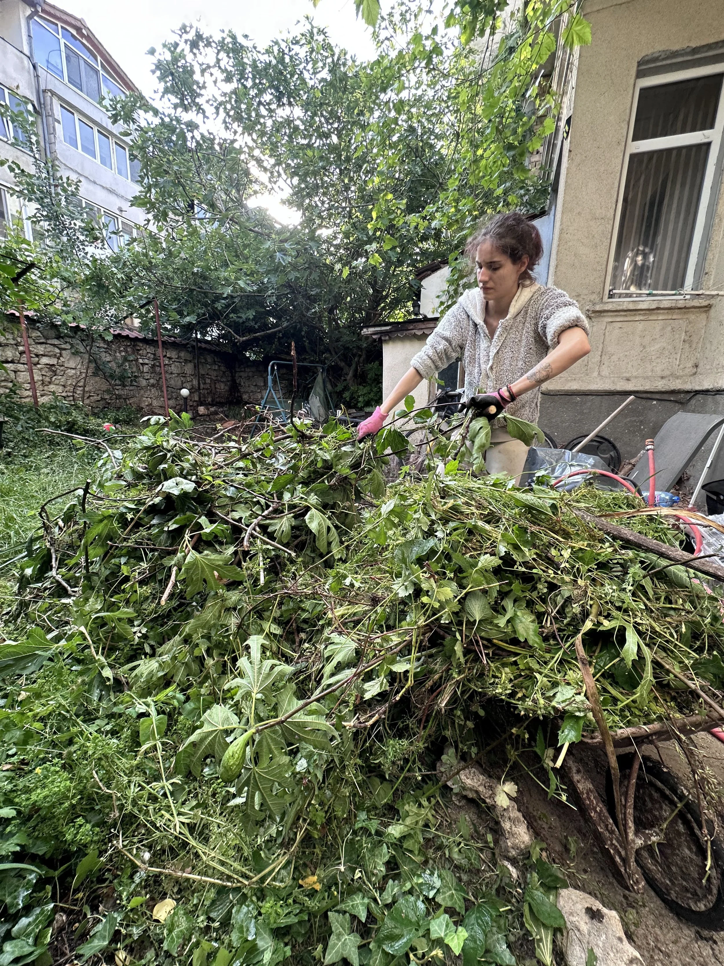 A woman wearing pink gloves collects garden waste, including branches and leaves, in a large pile in her backyard.