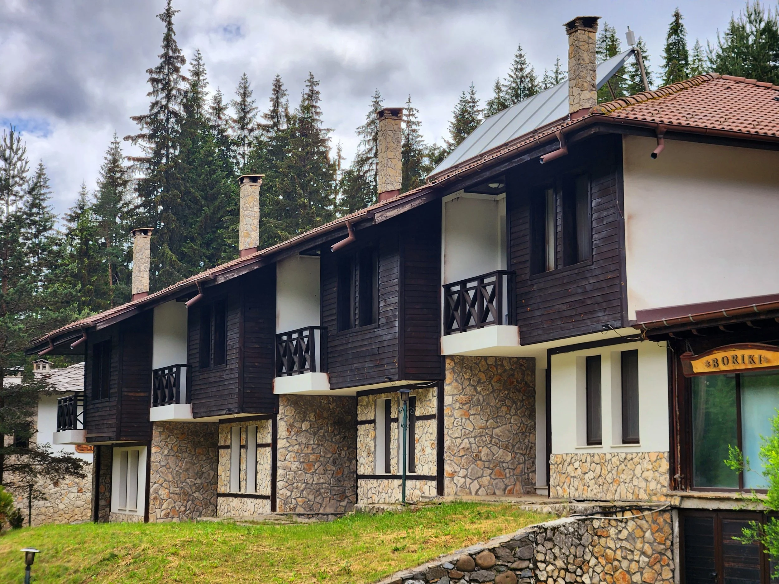 A multi-story house with a stone foundation, dark wooden siding, small balconies, and a sloped roof surrounded by tall evergreen trees.