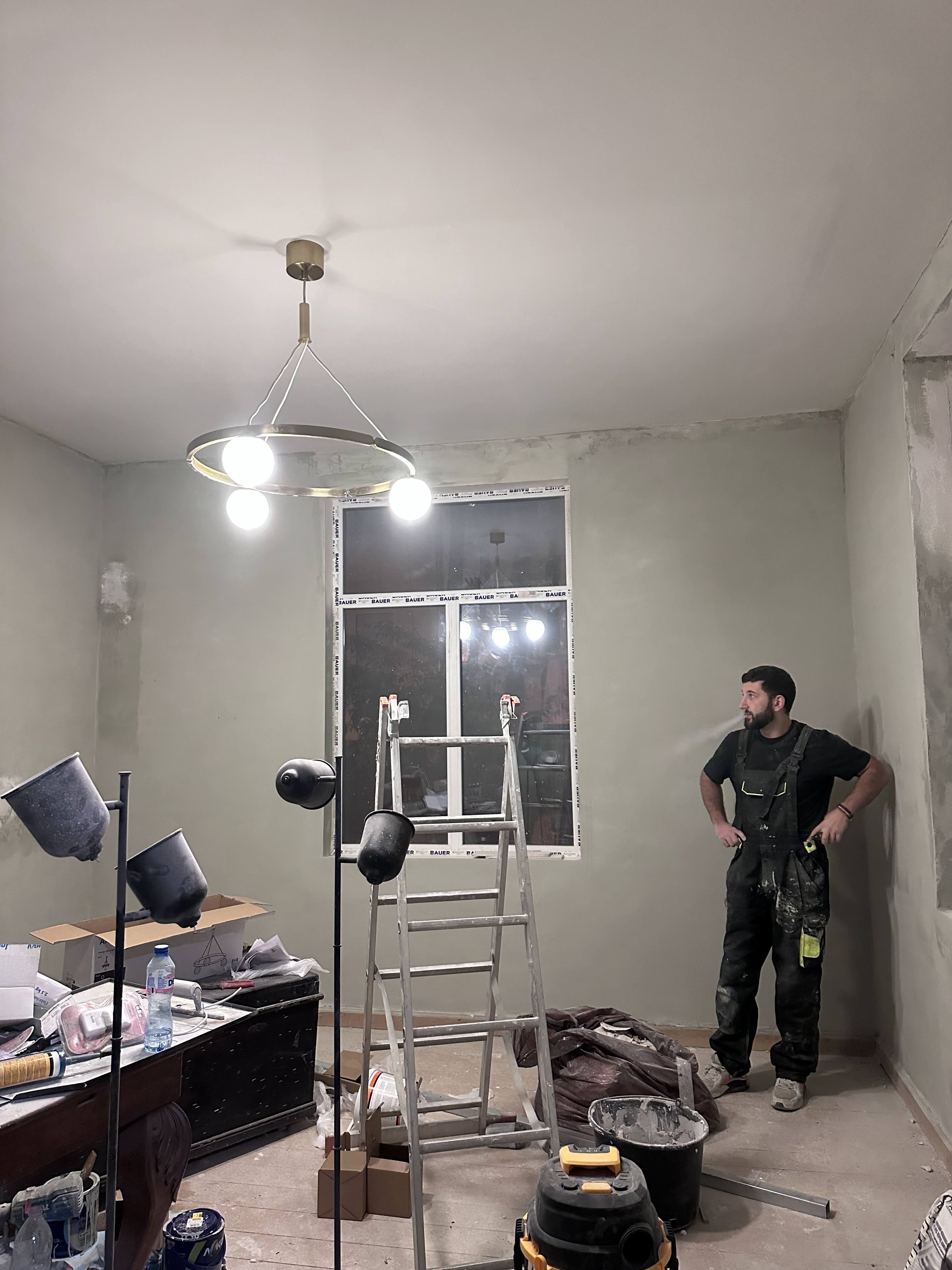 A man standing in a room under renovation with a ladder, construction tools, and furniture. The ceiling has a modern light fixture with three bulbs, and the walls are in the process of being painted or primed.