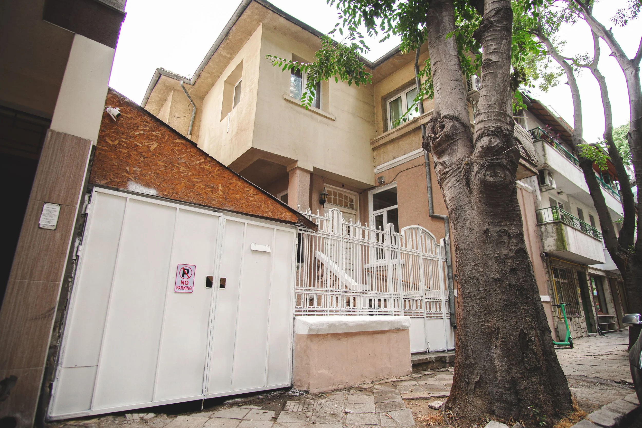 View of a multi-story residential building with a gated entrance, trees, and a scooter on the sidewalk.