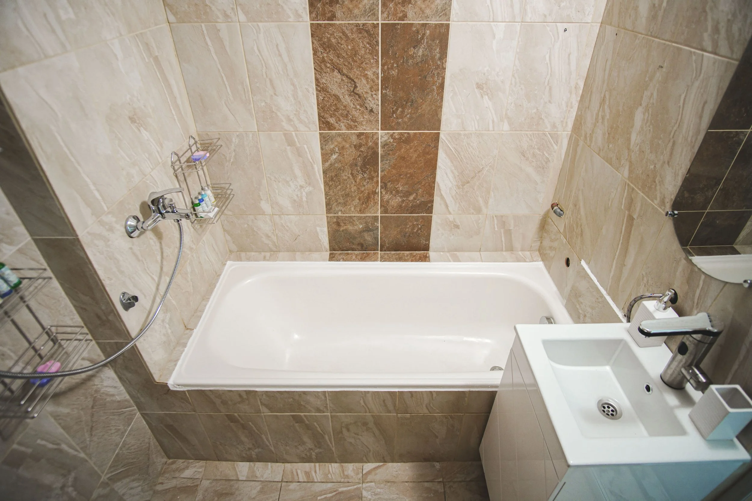 Bathroom with a bathtub, beige and brown tiled walls, a small white sink with a chrome faucet, and shower caddy with toiletries.