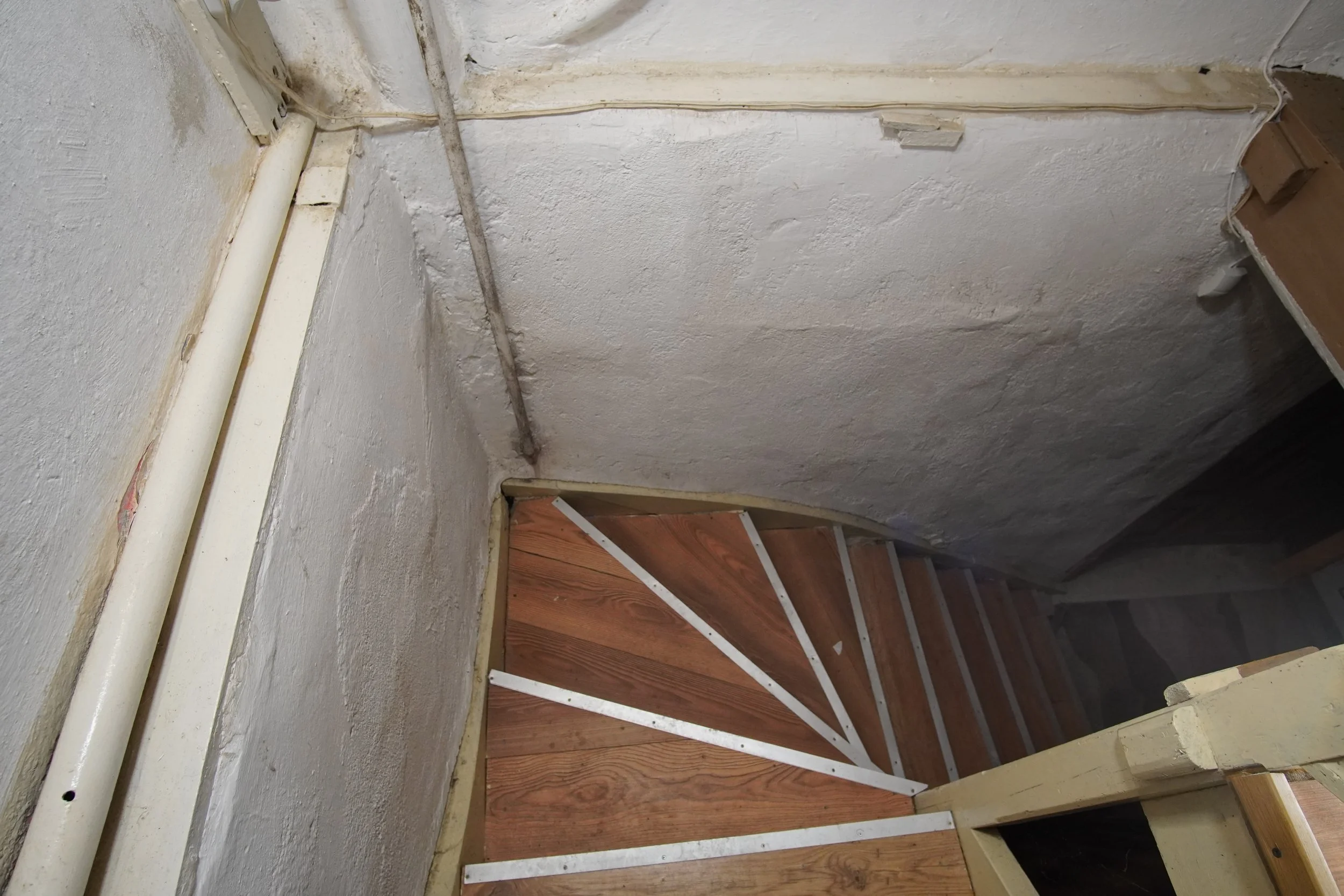 View of a small staircase under construction with wooden steps and metal brackets, adjacent to a textured white wall with a visible pipe and electrical wiring.