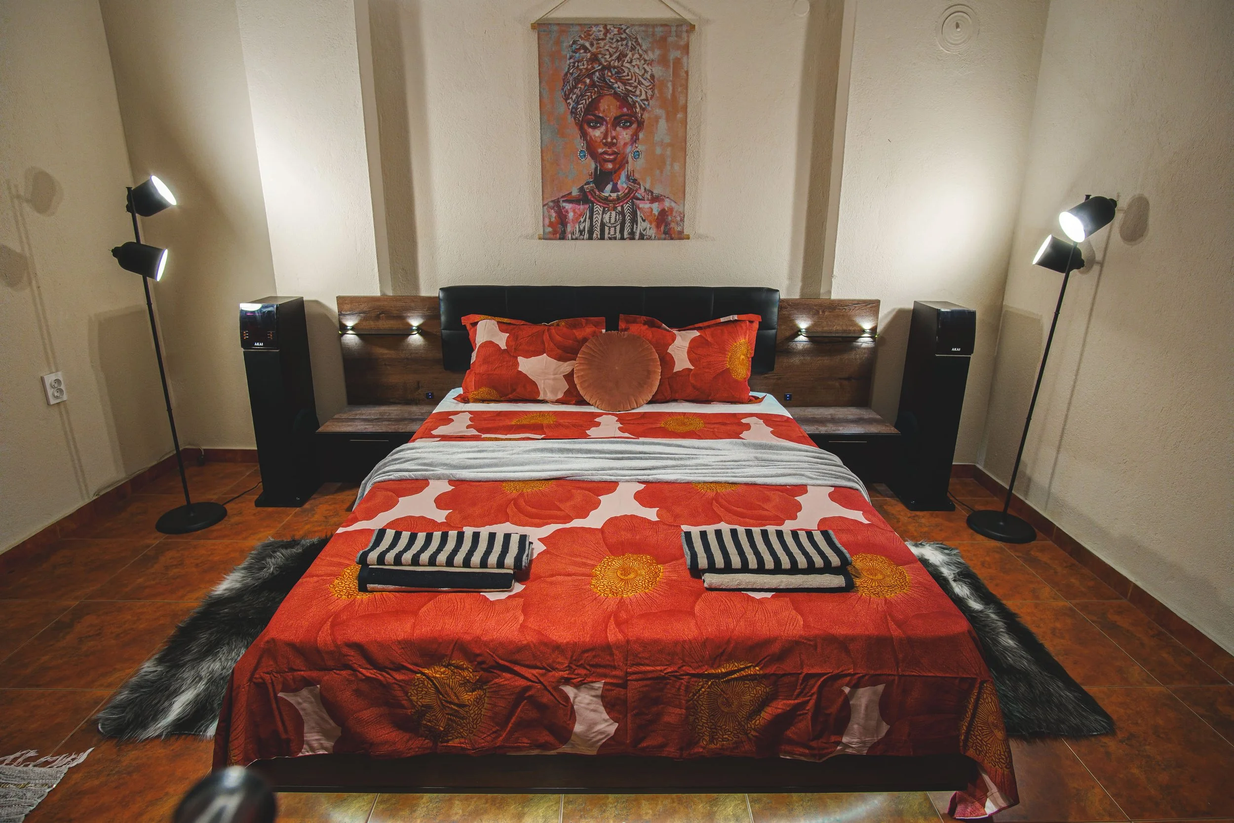 A cozy bedroom featuring a bed with orange floral bedding, orange pillows, and black and white striped towels, flanked by two black speakers, with two tall floor lamps on either side and a colorful portrait of a woman hanging on the wall above.