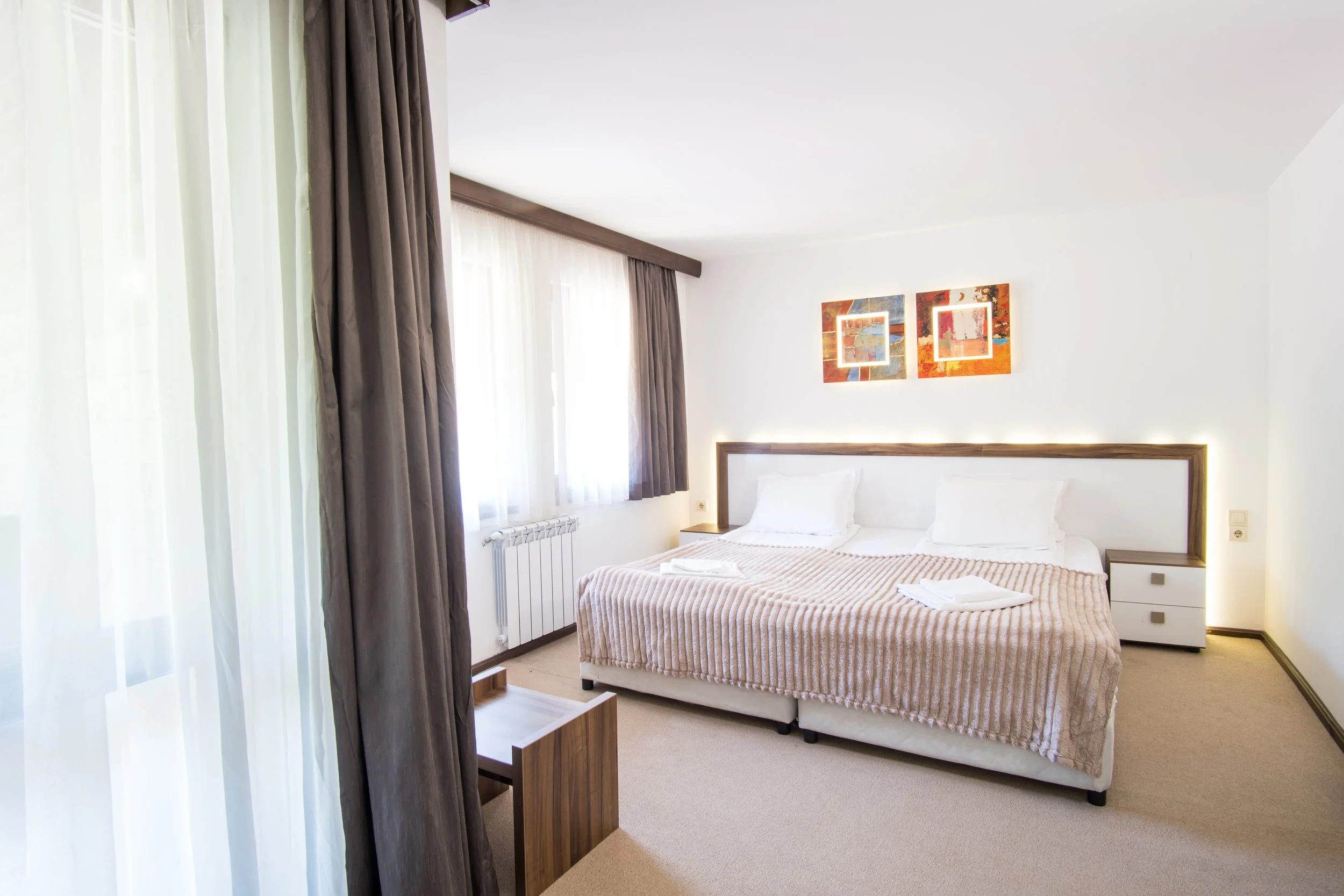 A clean, modern hotel room with a large bed, white bedding, and pillow, two abstract paintings above the headboard, a nightstand on each side, and windows with sheer white curtains and heavy brown drapes.