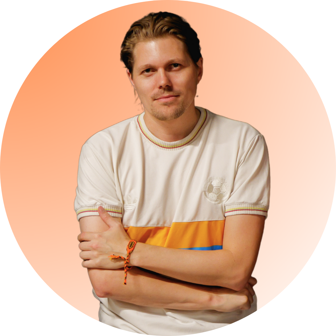 Man met blauwe ogen en bruin haar, gekleed in een wit en geel T-shirt met een voetballogo, staat met gekruiste armen voor een roze en oranje achtergrond.