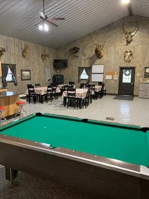 A game room with a pool table in the foreground, several tables and chairs, mounted animal heads and trophies on the wall, a mounted deer head, a television, windows, and door in the background.