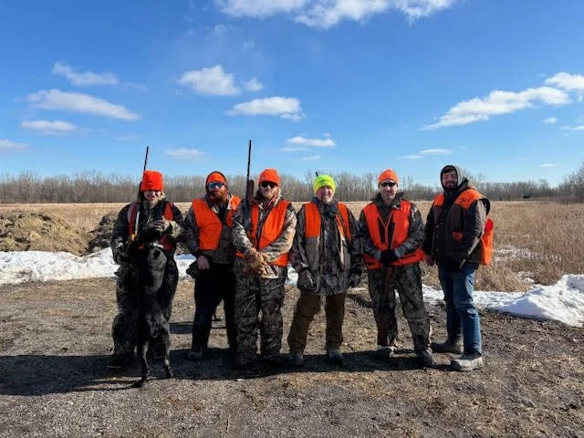 Six people and a dog standing outdoors in a line, dressed in hunting gear, on a clear day with snow patches and open field background.