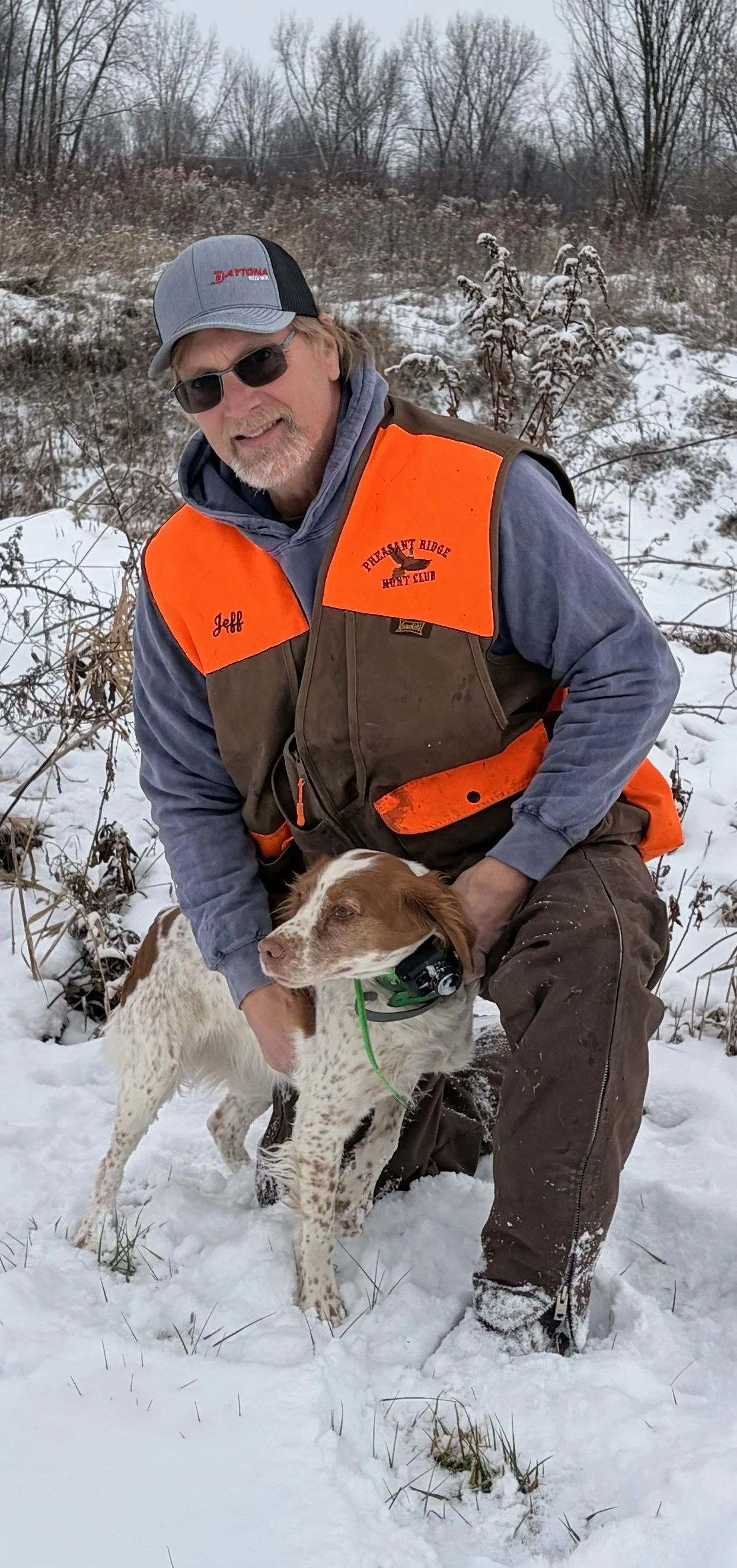 A man wearing sunglasses, an orange safety vest with the words "Pheasant Ridge Hunt Club" and the name "Jeff" on it, kneeling in snow with a dog. The dog has a collar with a green tag, and the background shows snow-covered ground and leafless trees.