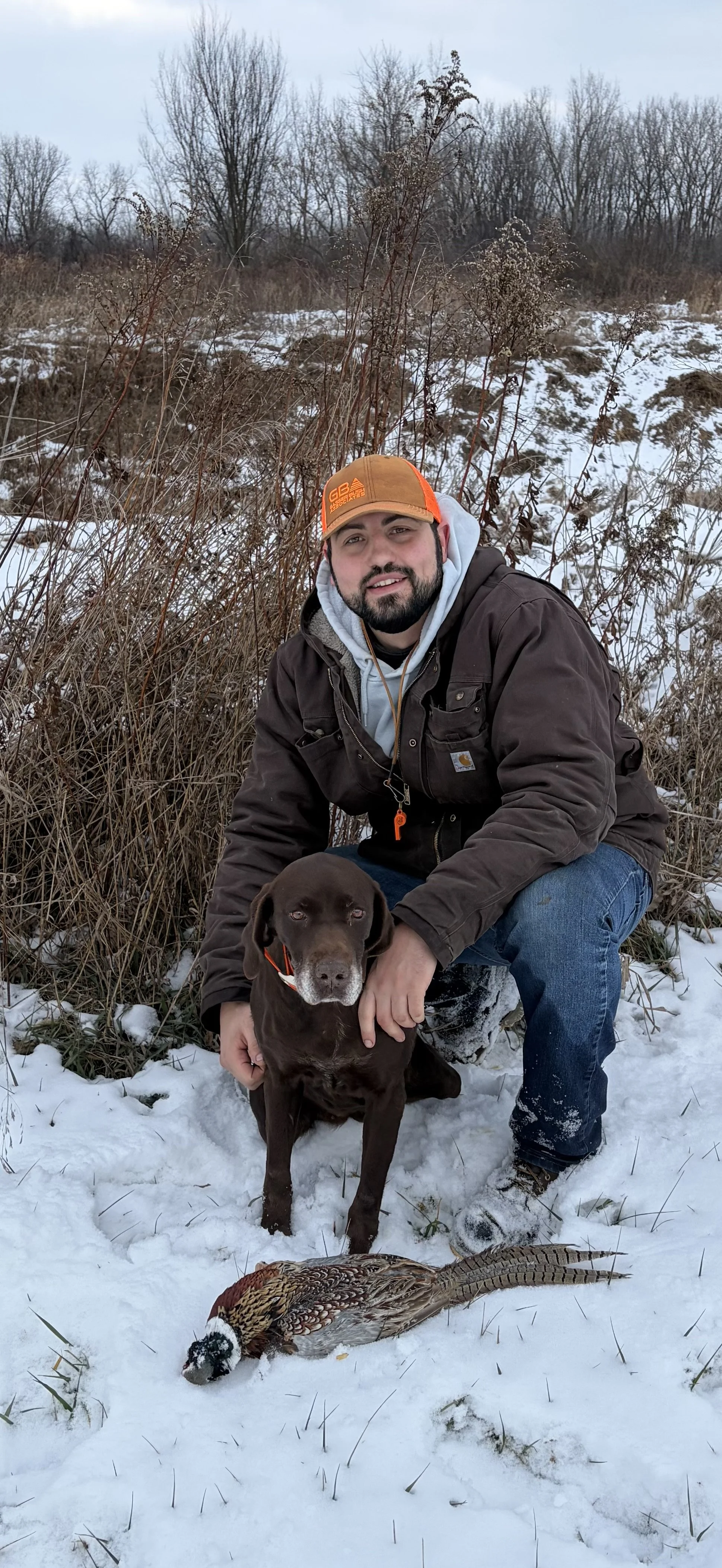 A man kneeling in the snow, holding a brown hunting dog, with a deceased pheasant on the ground in front of him. The background features snow-covered ground and leafless trees.