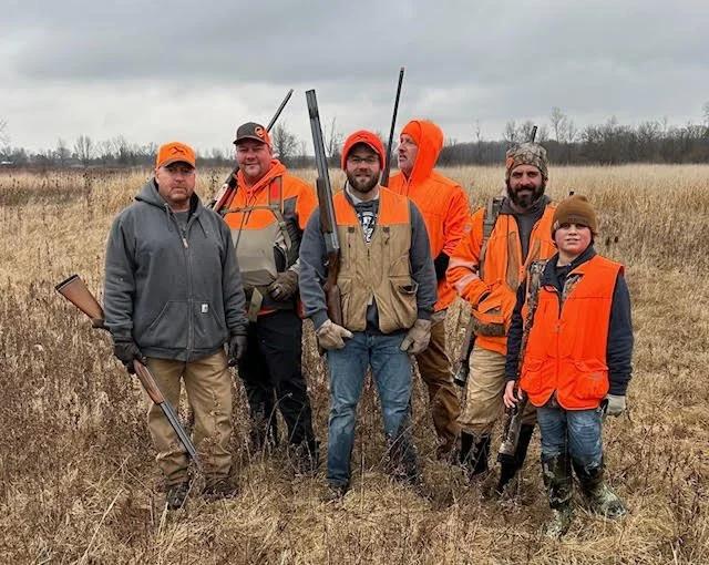 Six hunters wearing orange vests and jackets standing in a grassy field with overcast sky, holding shotguns.