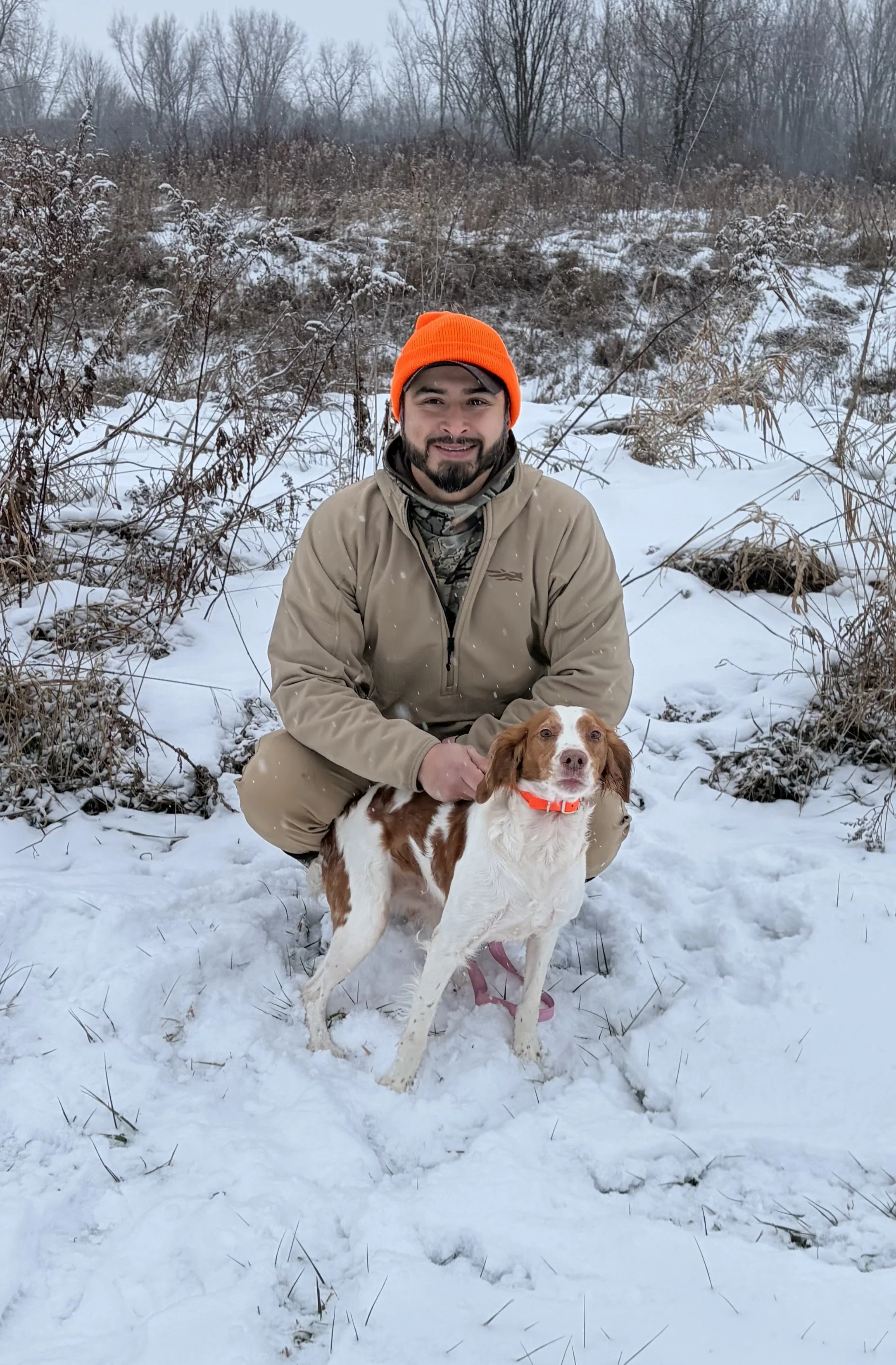 A man wearing a tan jacket and orange beanie squatting in the snow with a brown and white dog with a red collar outdoors in a snow-covered landscape with leafless trees and bushes.