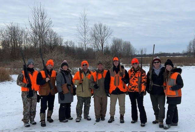 Group of people standing outdoors in snow wearing hunting vests and orange hats, some holding rifles.