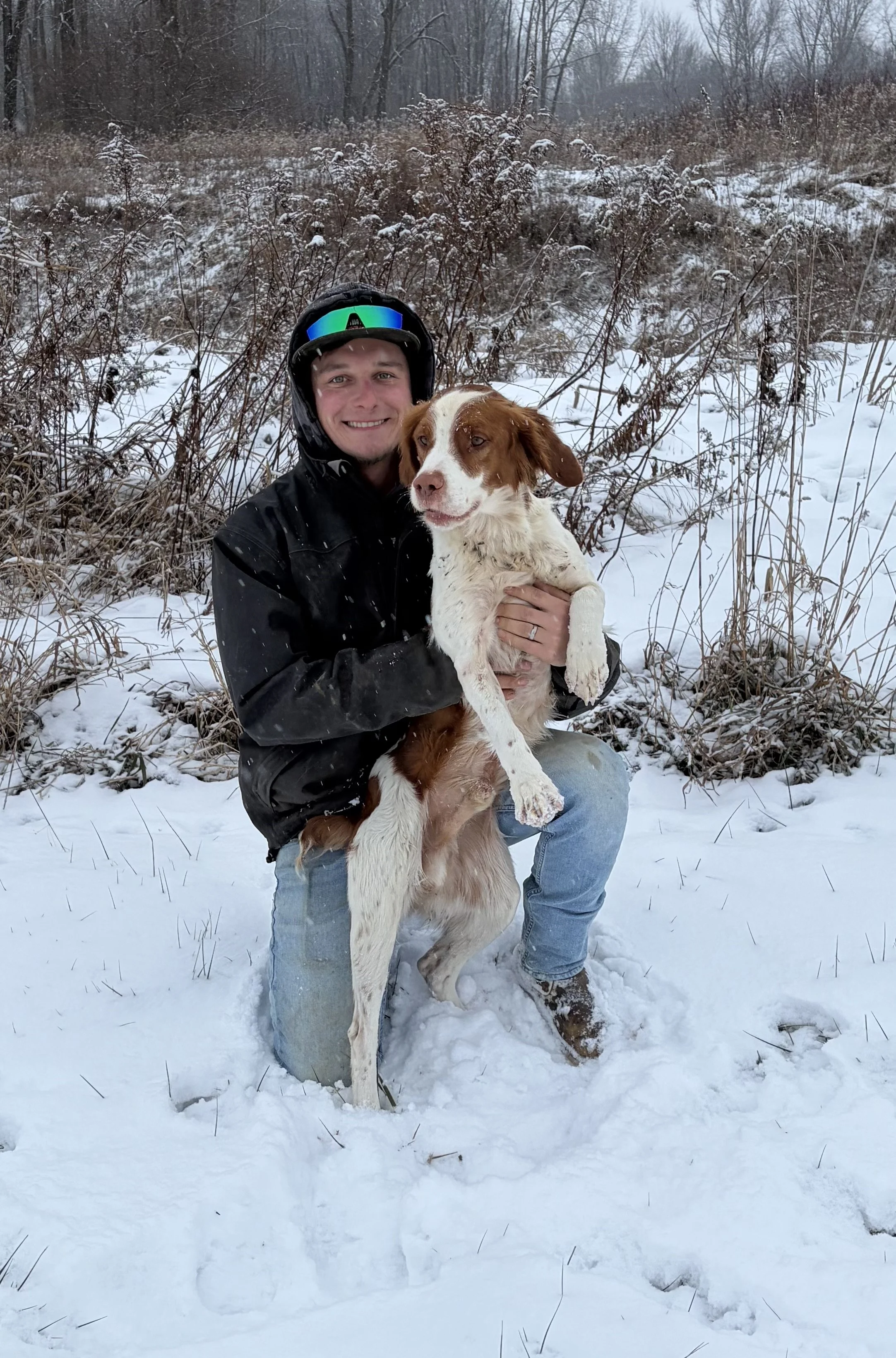 A man in winter clothing and a helmet kneeling in snow, smiling and holding a large brown and white dog in a snowy outdoor setting with bushes and trees.