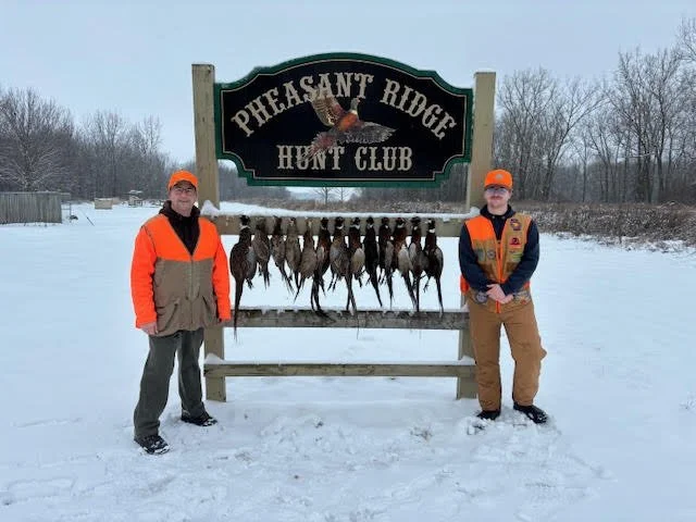 Two people in hunting gear stand outdoors in snow next to a sign that reads 'Pheasant Ridge Hunt Club' with a row of pheasant birds hanging from it.