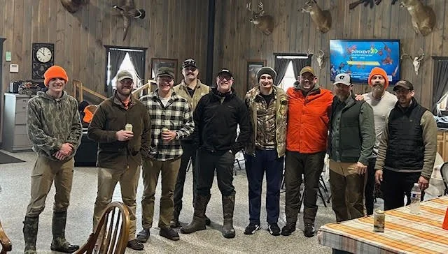 Group of men standing together indoors, some holding drinks, with rustic decor and mounted animal heads on the wall.