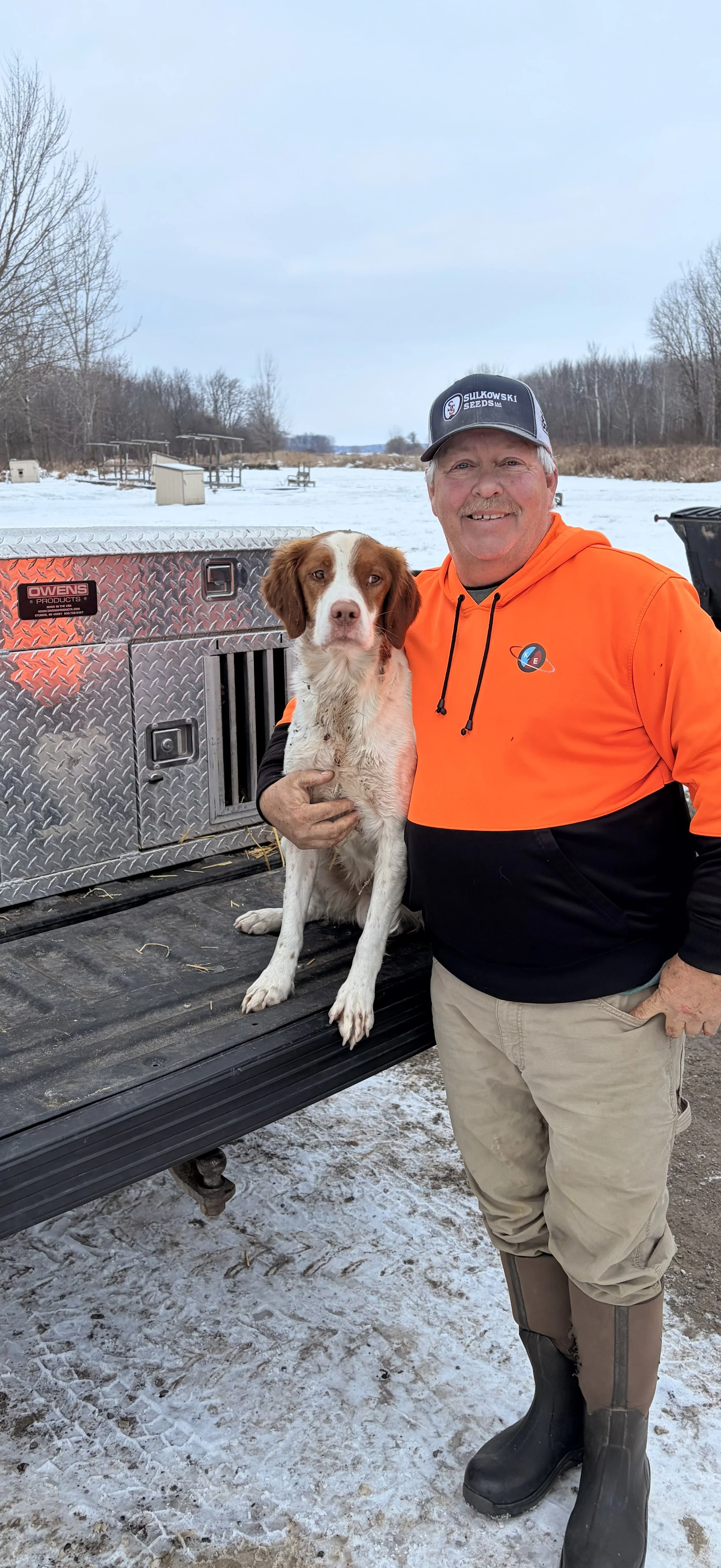 A man wearing an orange hoodie and black cap holding a brown and white dog on a truck bed outdoors with snow and leafless trees in the background.