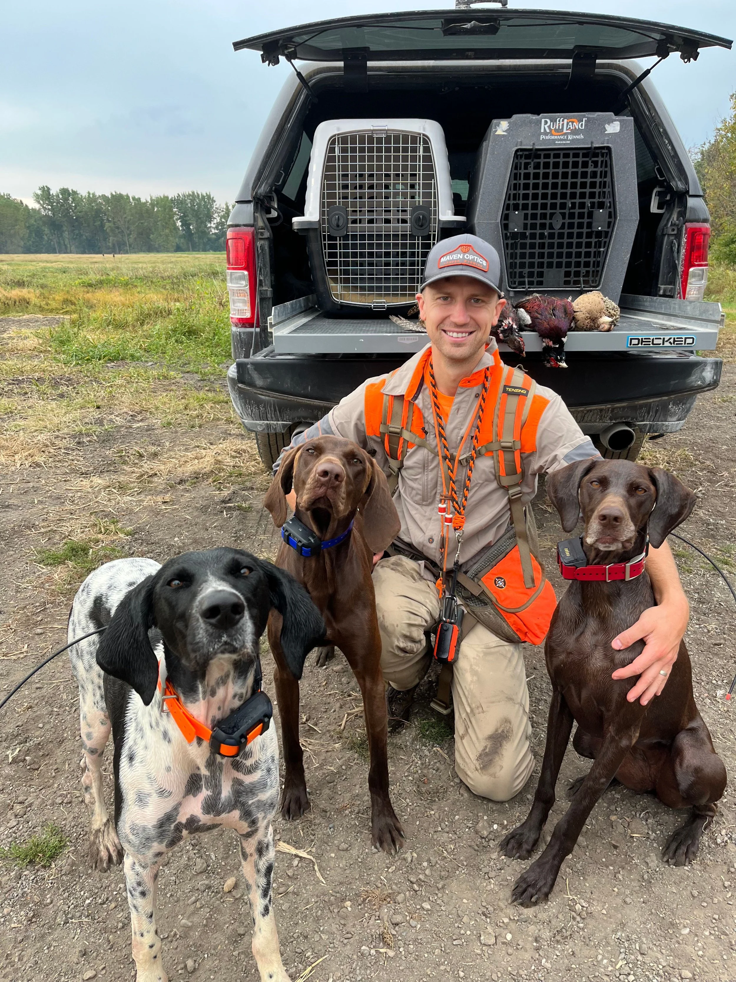 A man kneeling outdoors in front of a black pickup truck with an open tailgate. He is smiling and surrounded by three dogs, two of which are sitting on the ground and one is standing. Behind him are two large dog crates, one white and one black. The dogs are wearing orange collars and are likely hunting dogs. The man is dressed in outdoor clothing and has hunting gear around his neck.